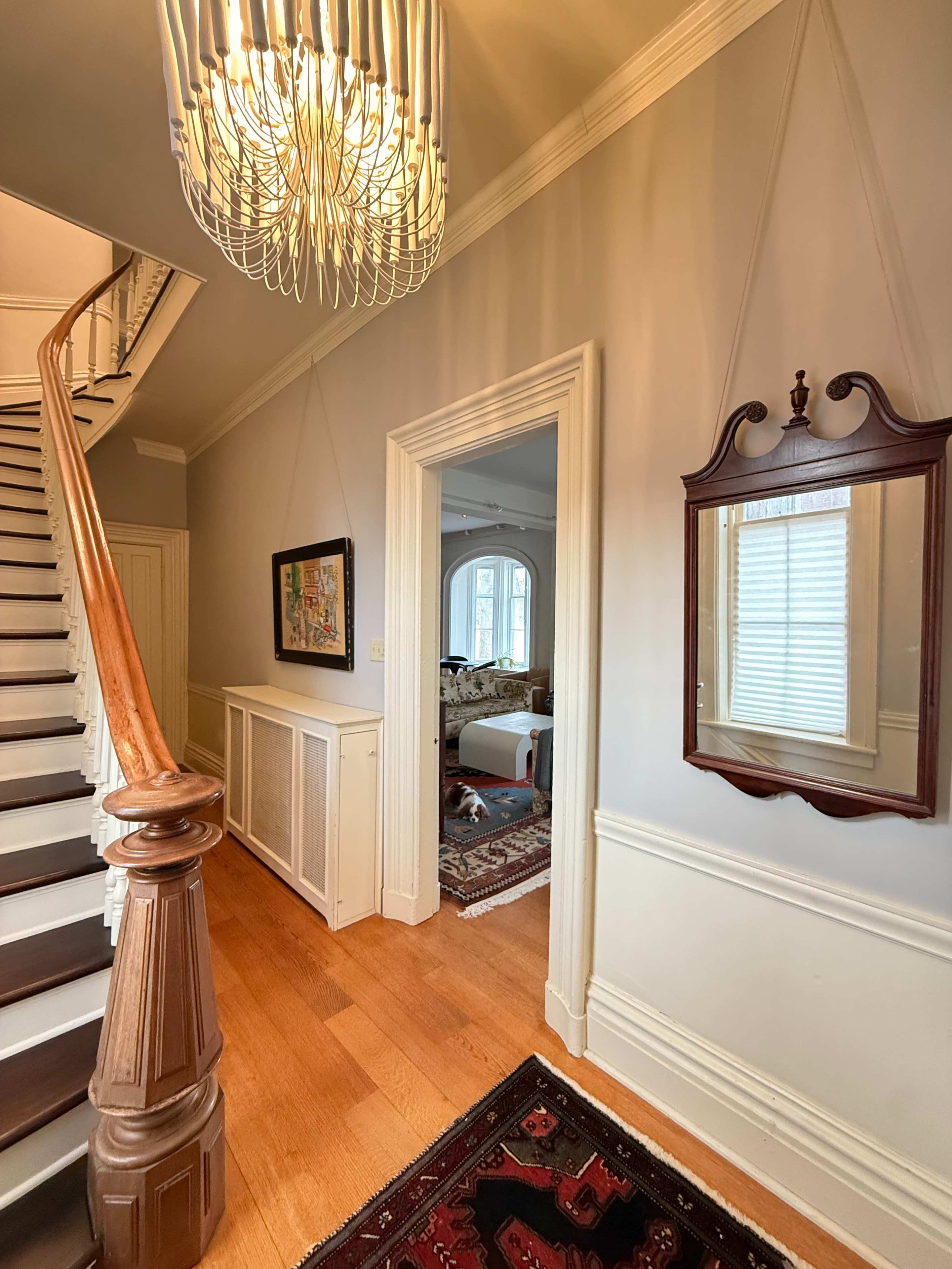 The image shows a staircase leading to an upper floor, with wooden railings, a decorative mirror on the wall, and a view into a room with a plush rug and furnishings.