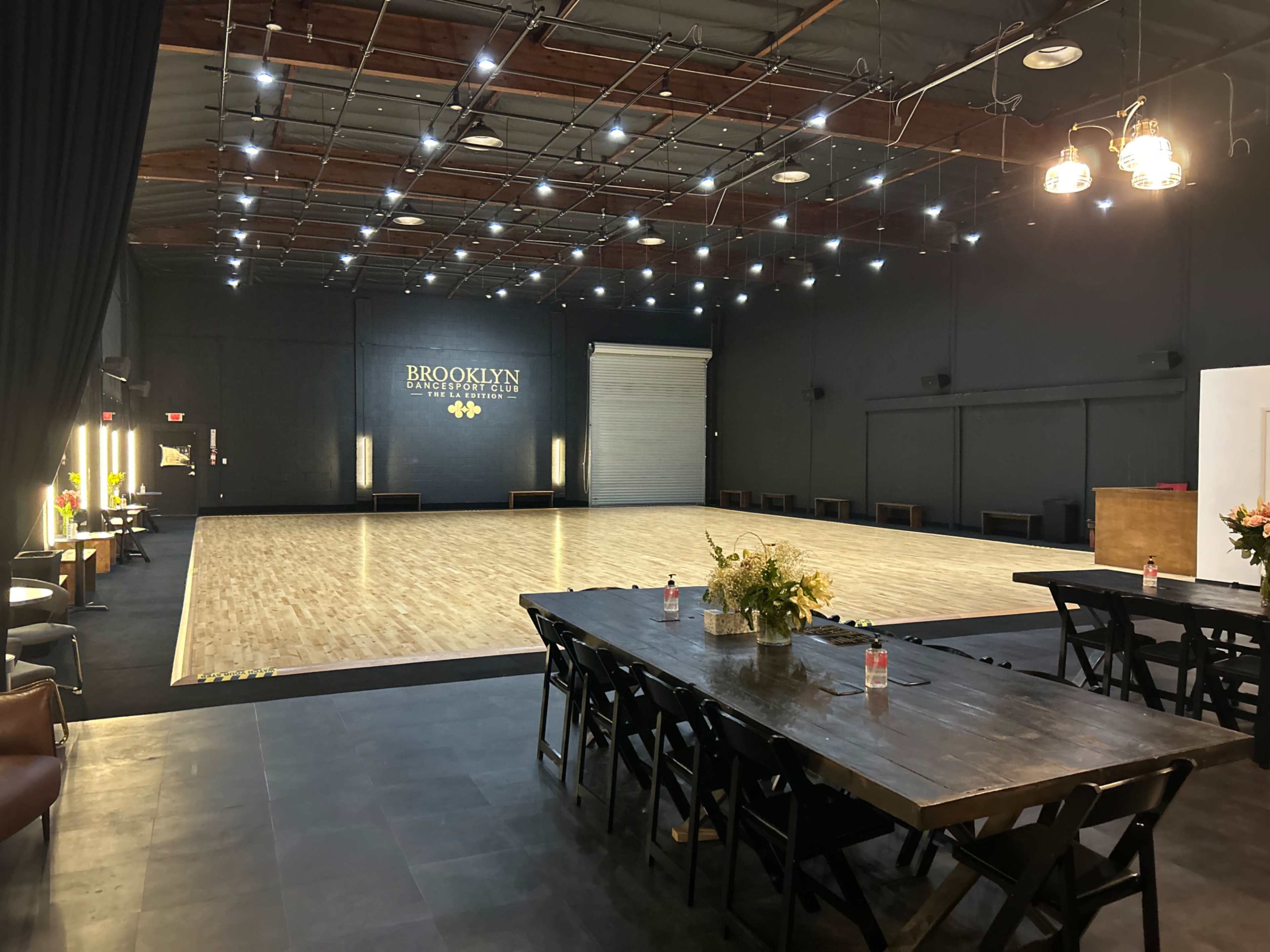A spacious dance studio with a polished wooden floor, dim lighting, and a long table set for dining or gatherings along one side.