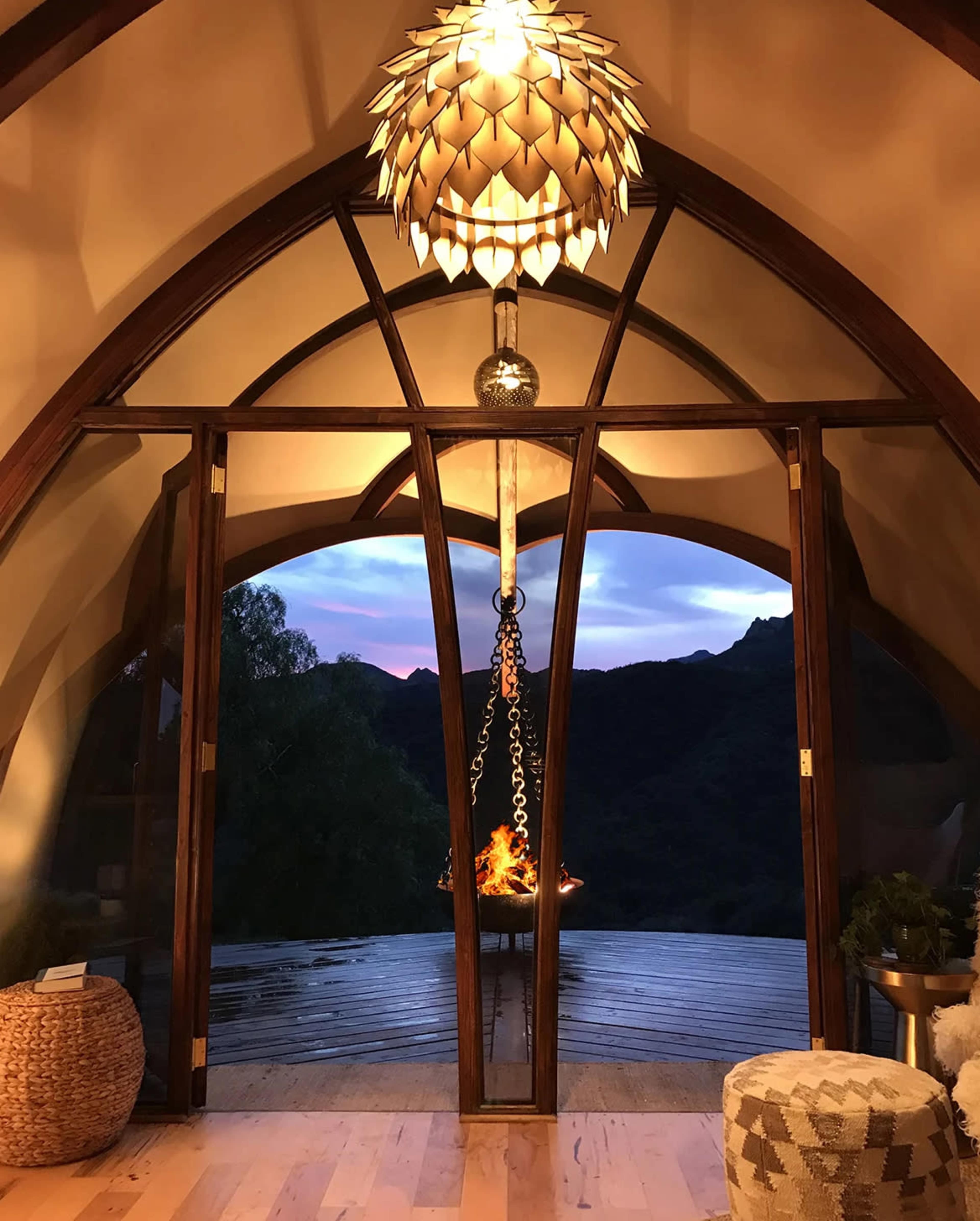 A modern interior features large glass doors opening to a scenic view of mountains at sunset, with a unique pendant light and a hanging fire feature.