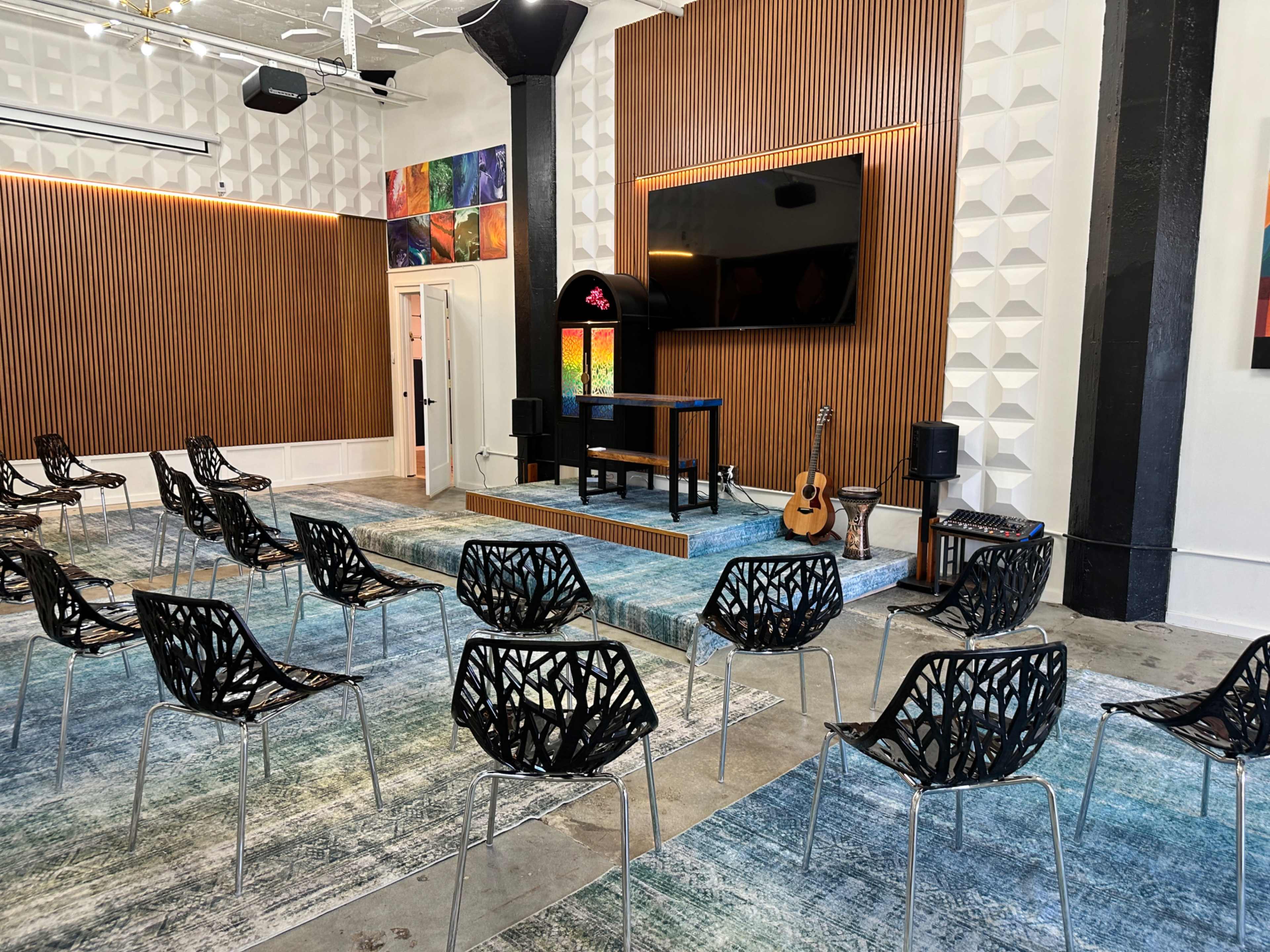 The image shows a seminar room with rows of black chairs arranged on a colorful carpet, featuring a stage with a guitar and a large screen mounted on a wooden panel wall.