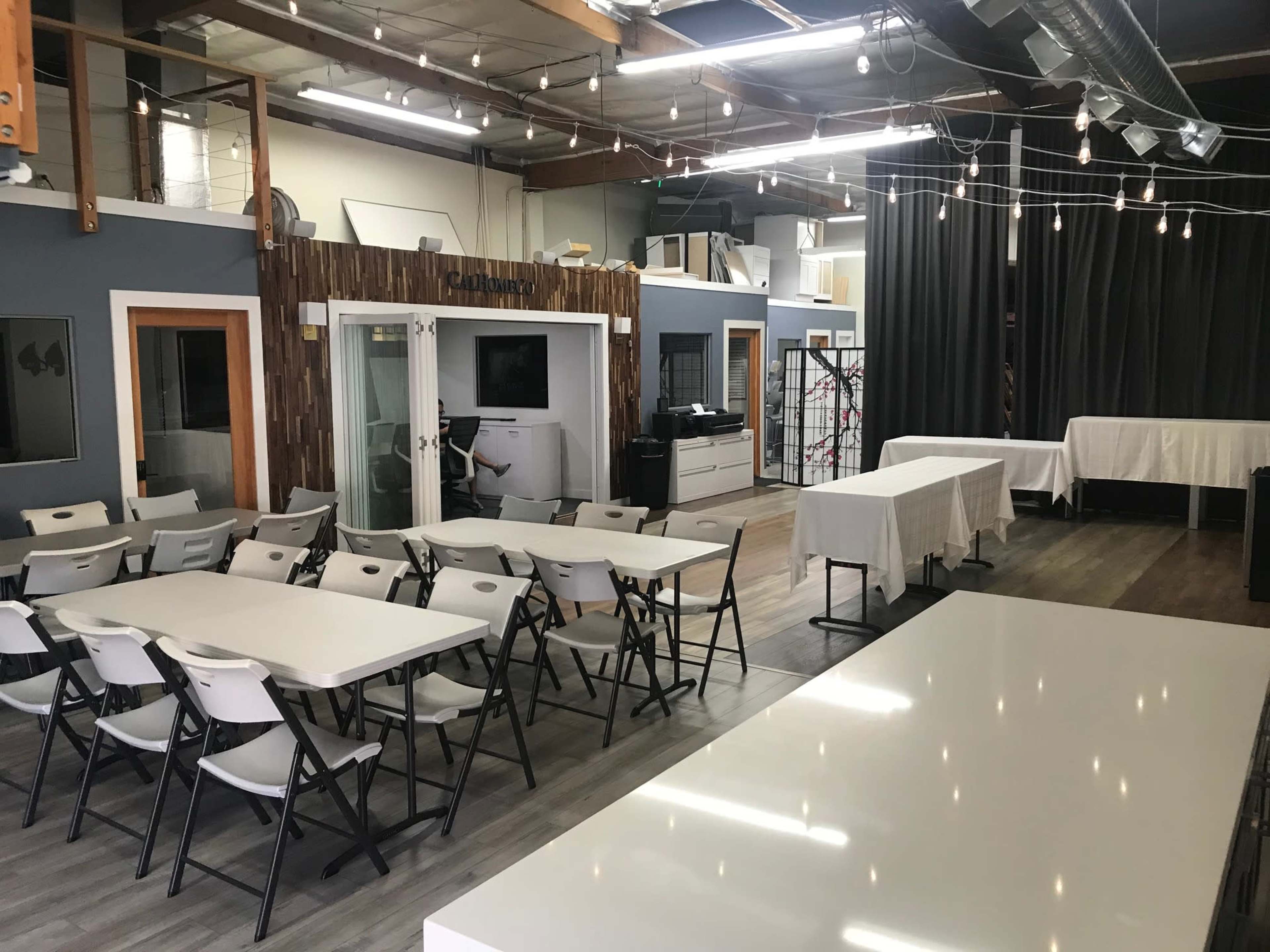 The image shows a spacious indoor area with folding chairs arranged in rows and several tables set up, surrounded by walls adorned with artwork and curtains.