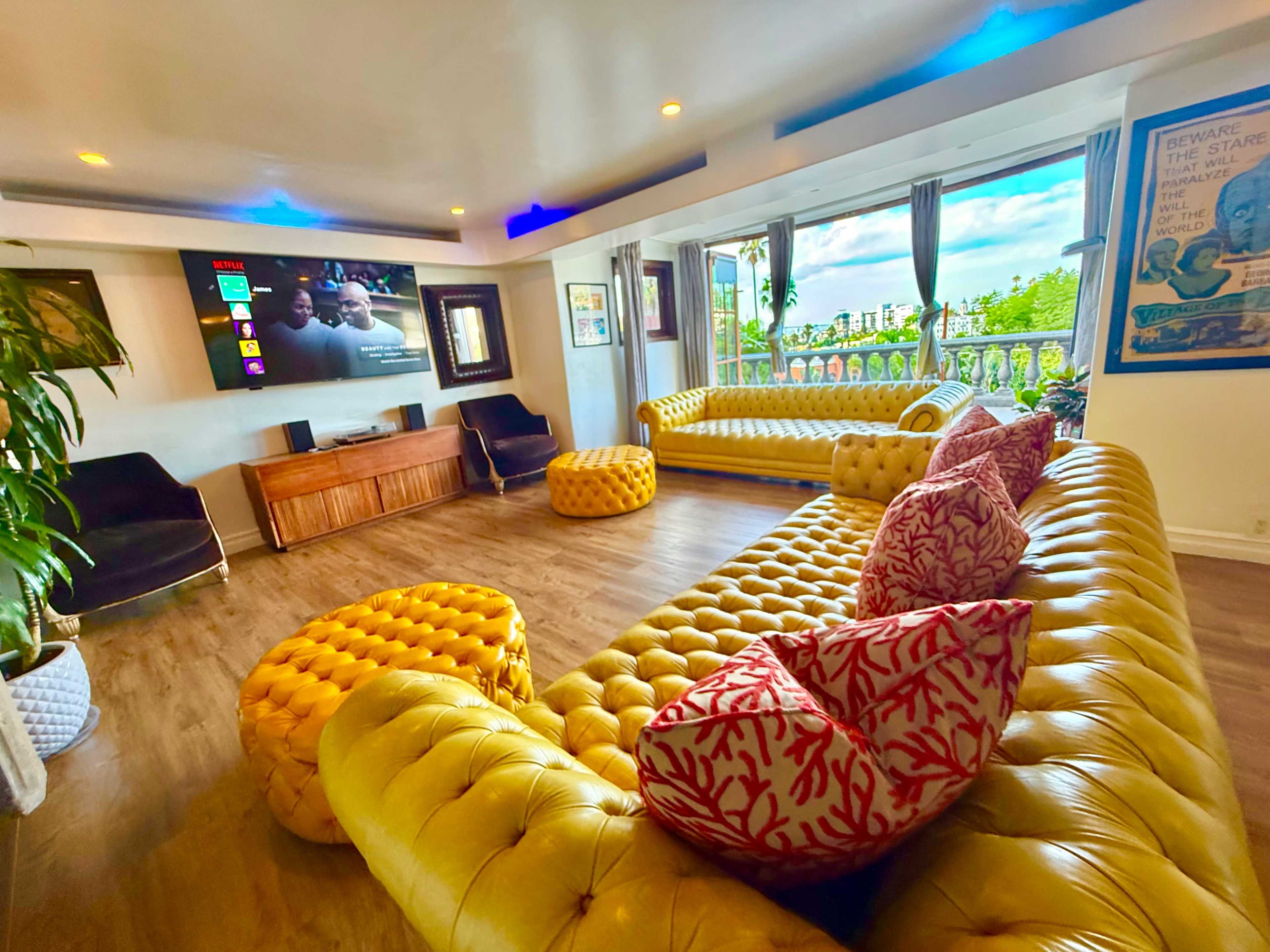 The living room features yellow tufted sofas, colorful cushions, a wooden entertainment unit, and large windows showcasing a lush outdoor view.