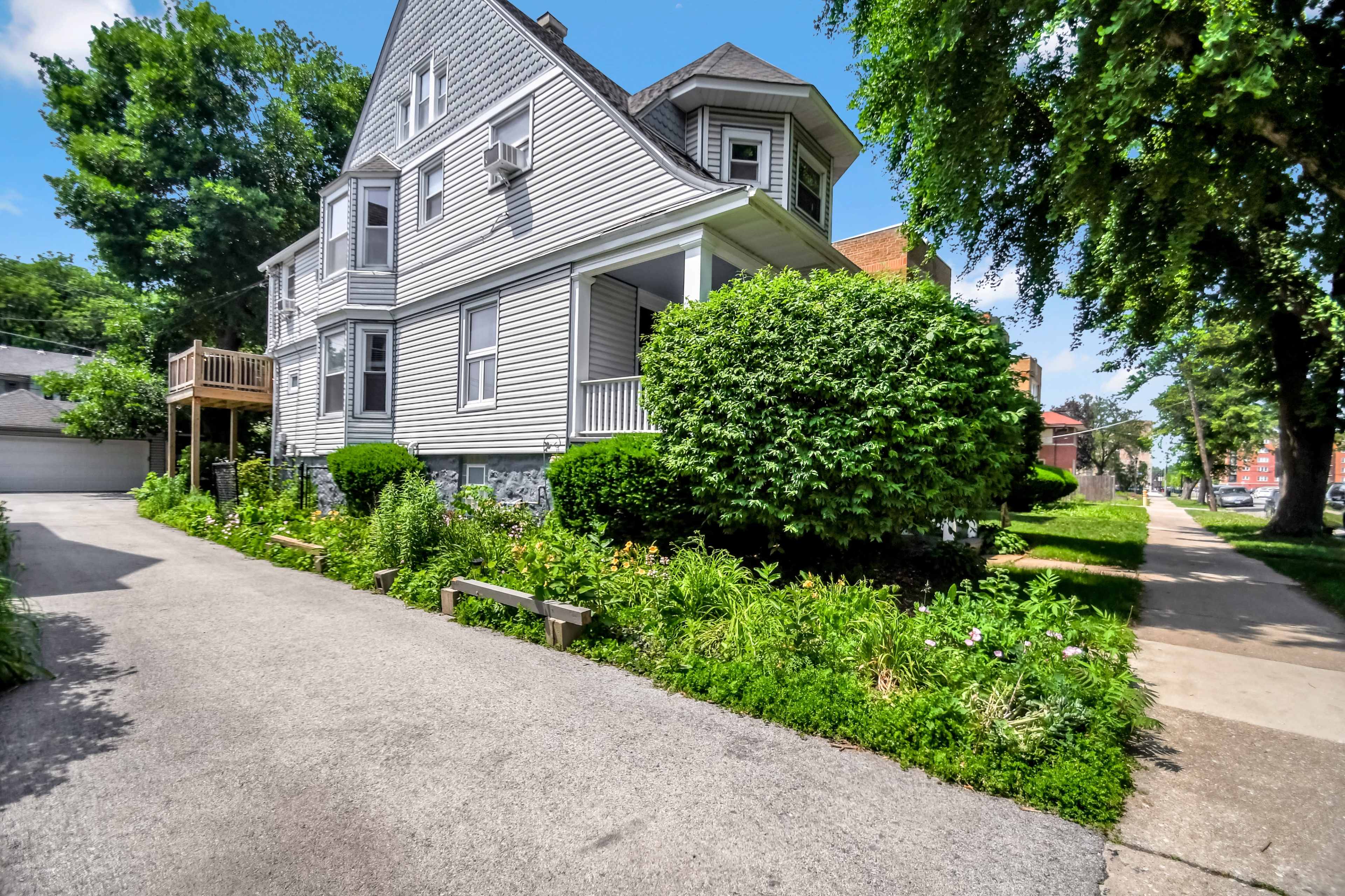 A gray house with a steep roof and porch is situated next to a well-maintained garden and walkway in a residential neighborhood.