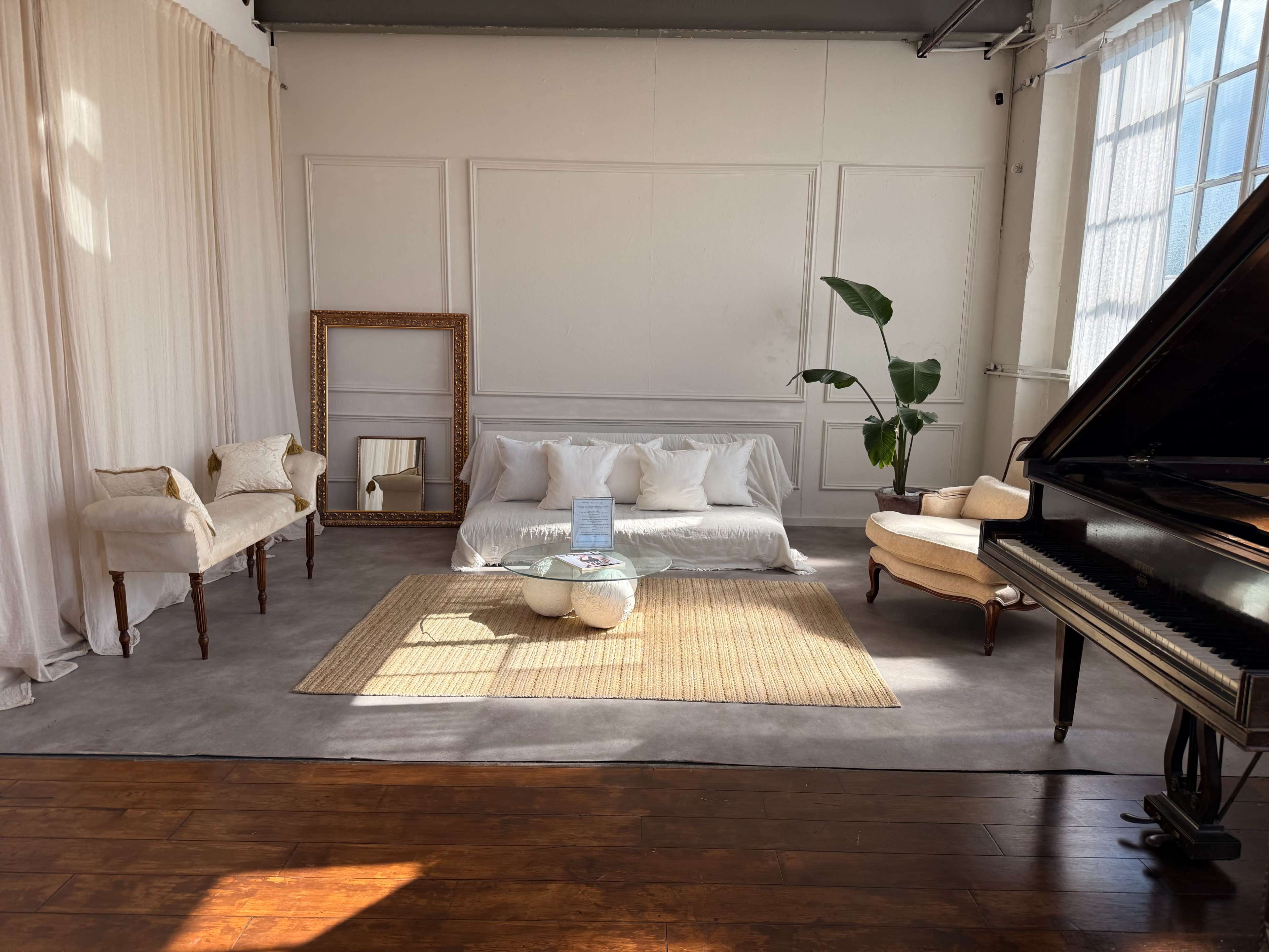 The image shows a bright, minimalist living space with a sofa, a glass coffee table, a piano, and decorative plants against a backdrop of large windows and neutral-colored walls.