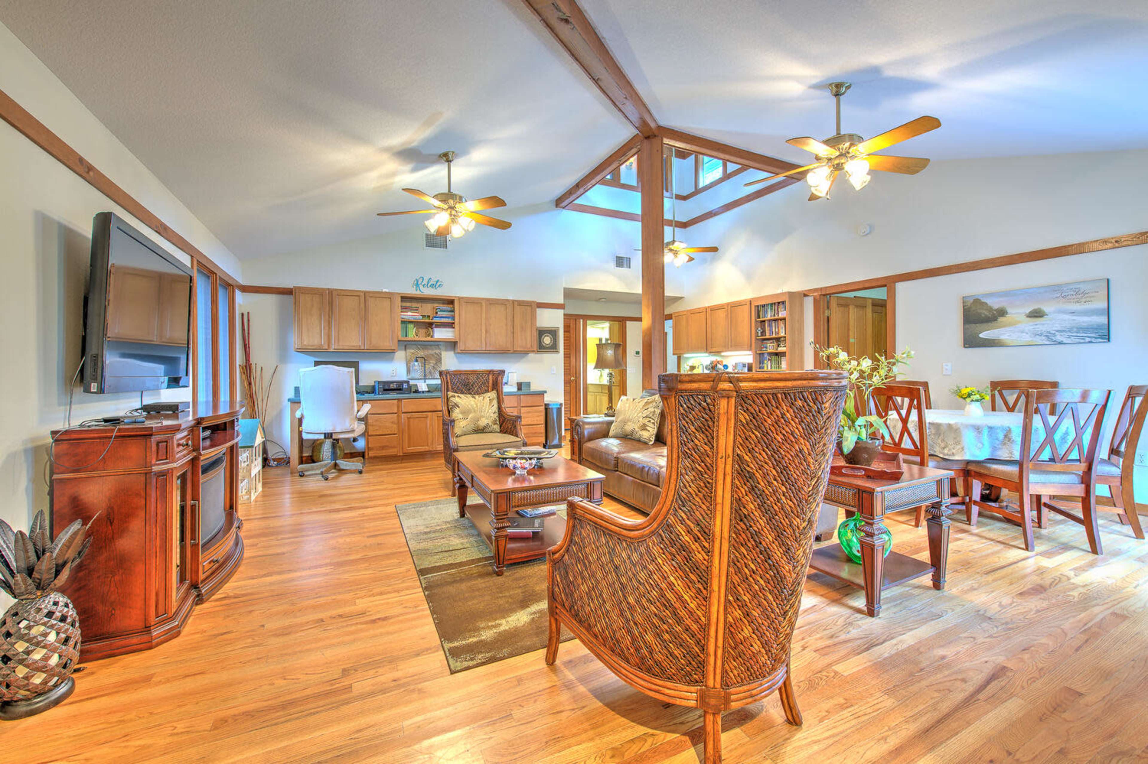 A spacious living area with wooden flooring, a seating arrangement featuring a couch and chairs, and a dining table, all illuminated by ceiling fans and natural light from large windows.