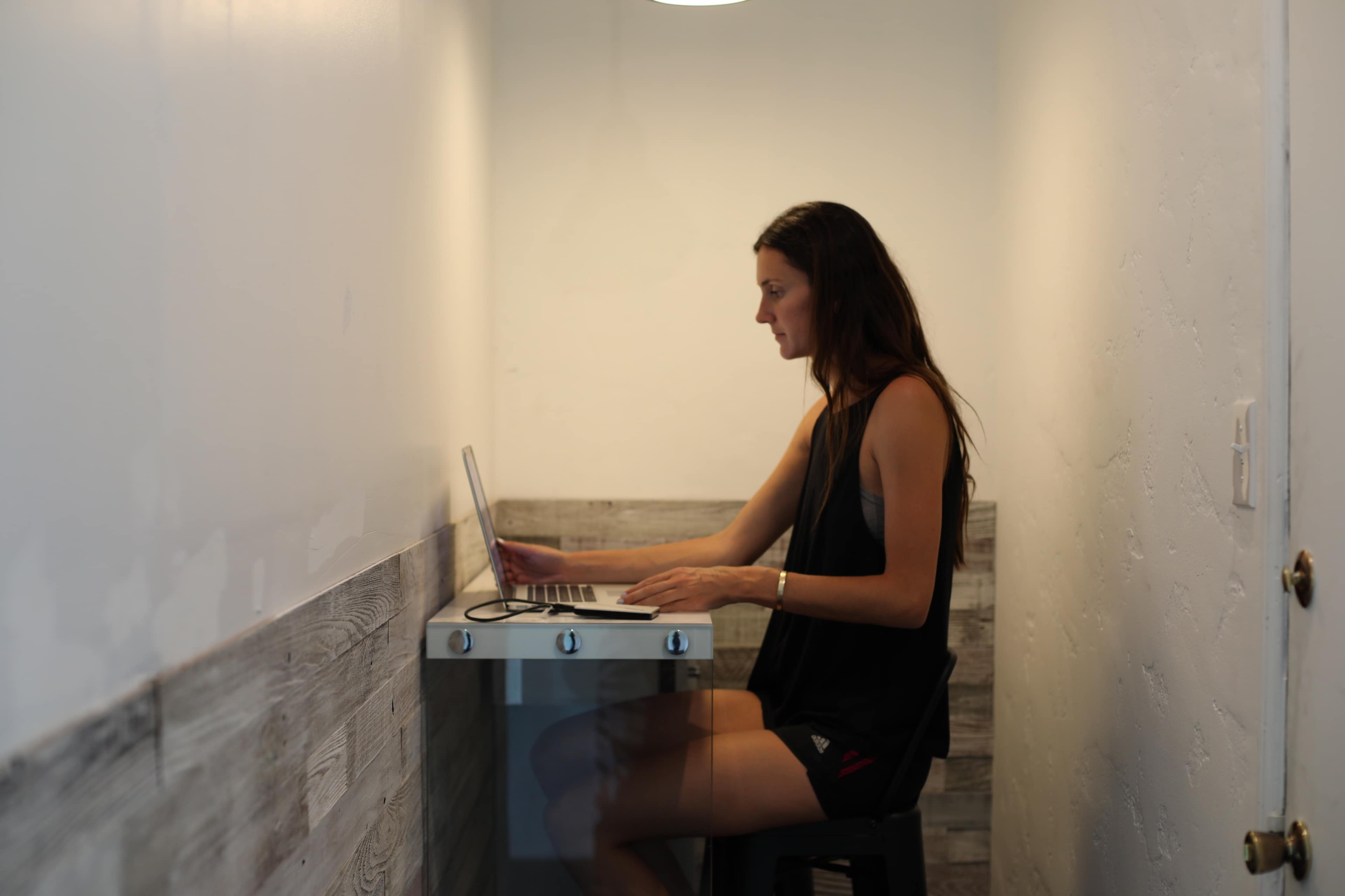 A woman sits at a small desk in a narrow room, using a laptop with a pendant light overhead.