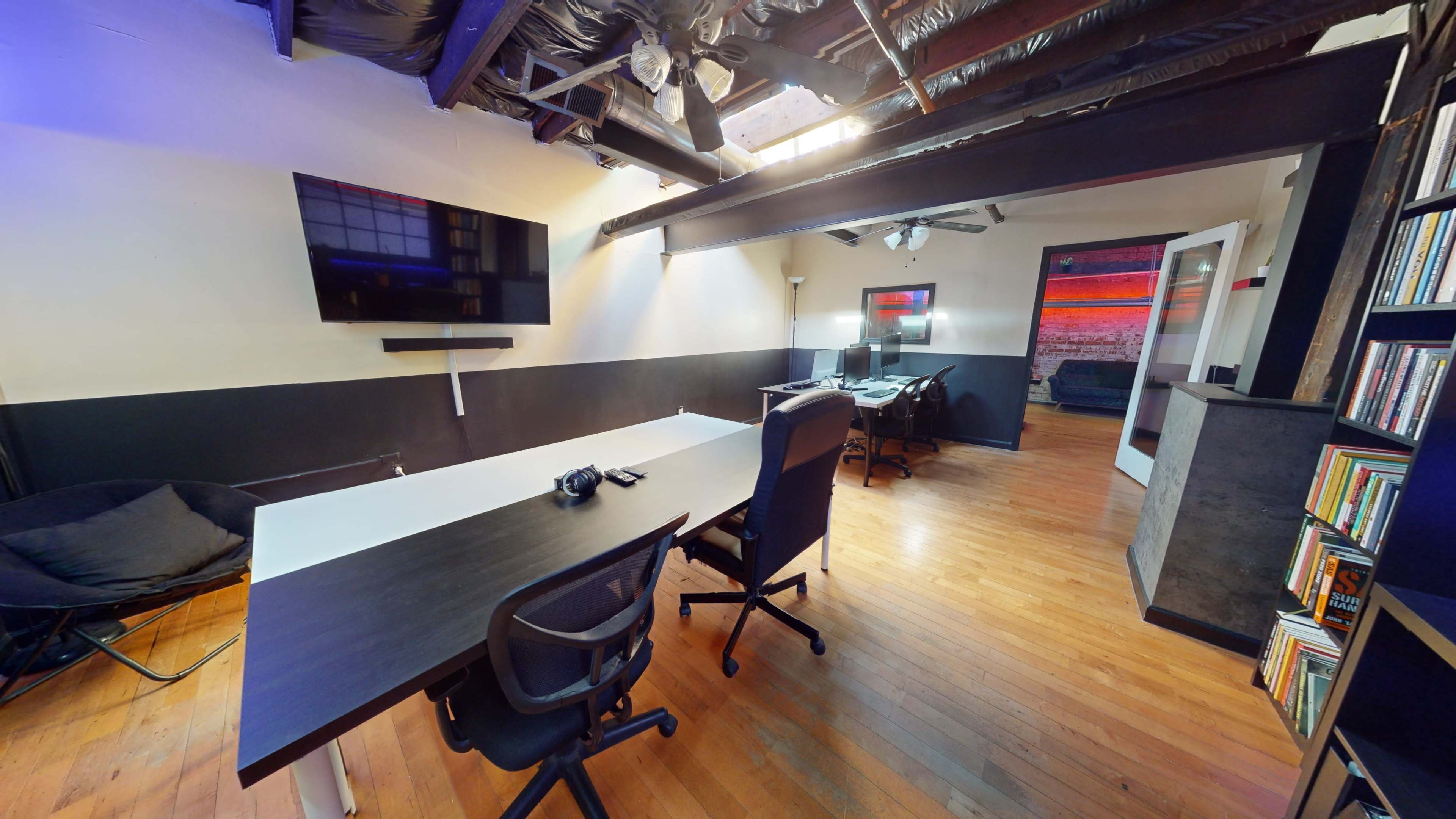 A modern office space featuring a large table, black chairs, and two workstations, with exposed beams and wooden flooring.