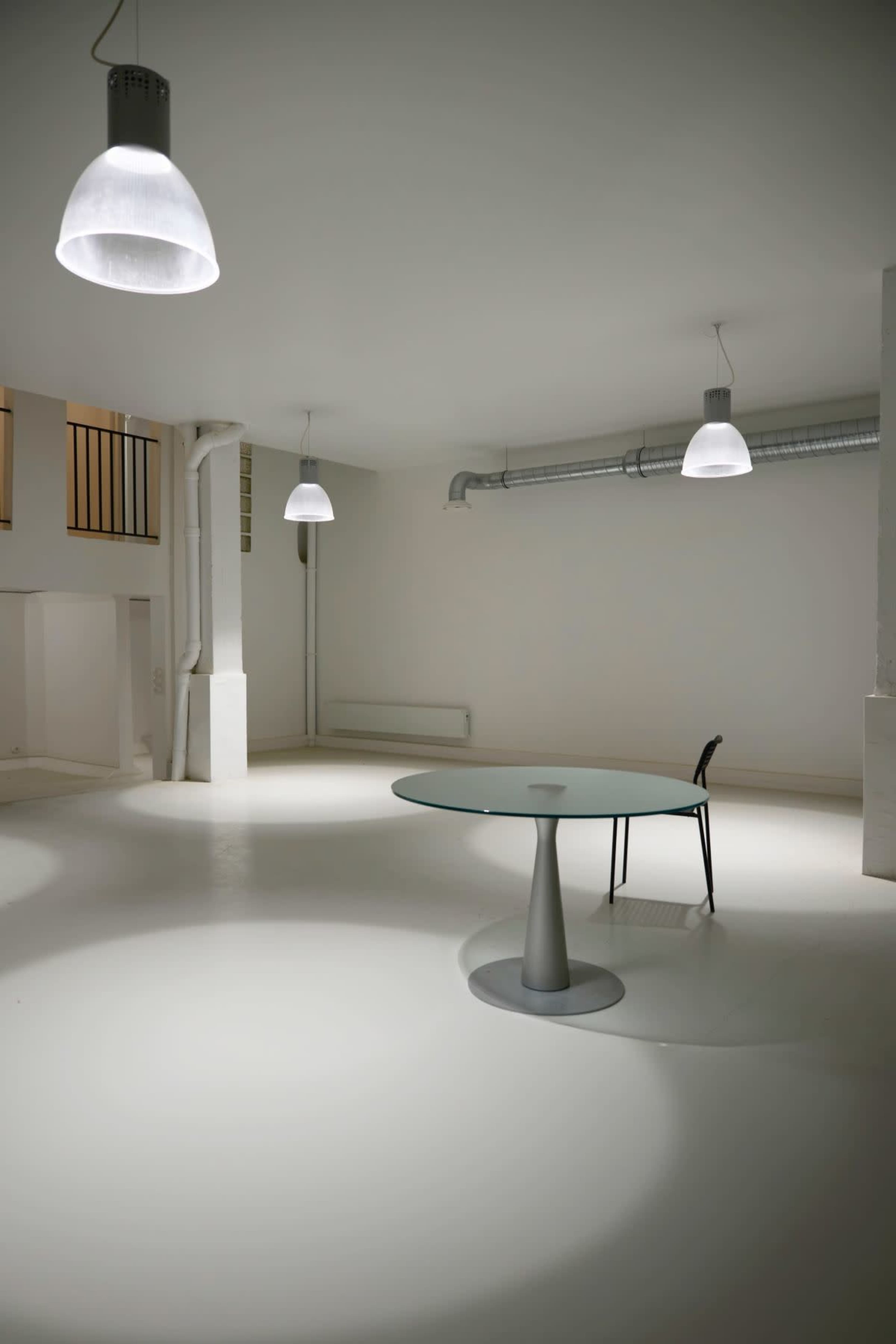 The image shows a minimalistic, well-lit interior space featuring a round glass table and a single black chair.