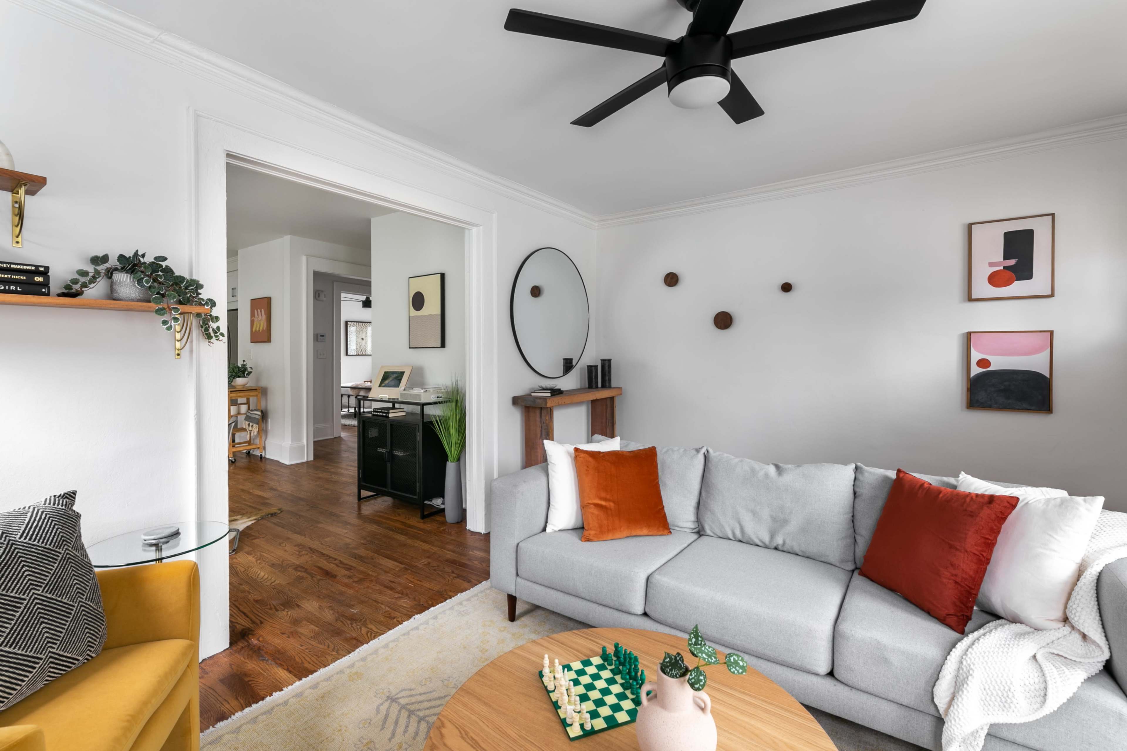 A modern living room features a light gray sofa with orange and white pillows, a round wooden coffee table with a chess set, and a fan overhead, while a doorway leads to an adjoining room.