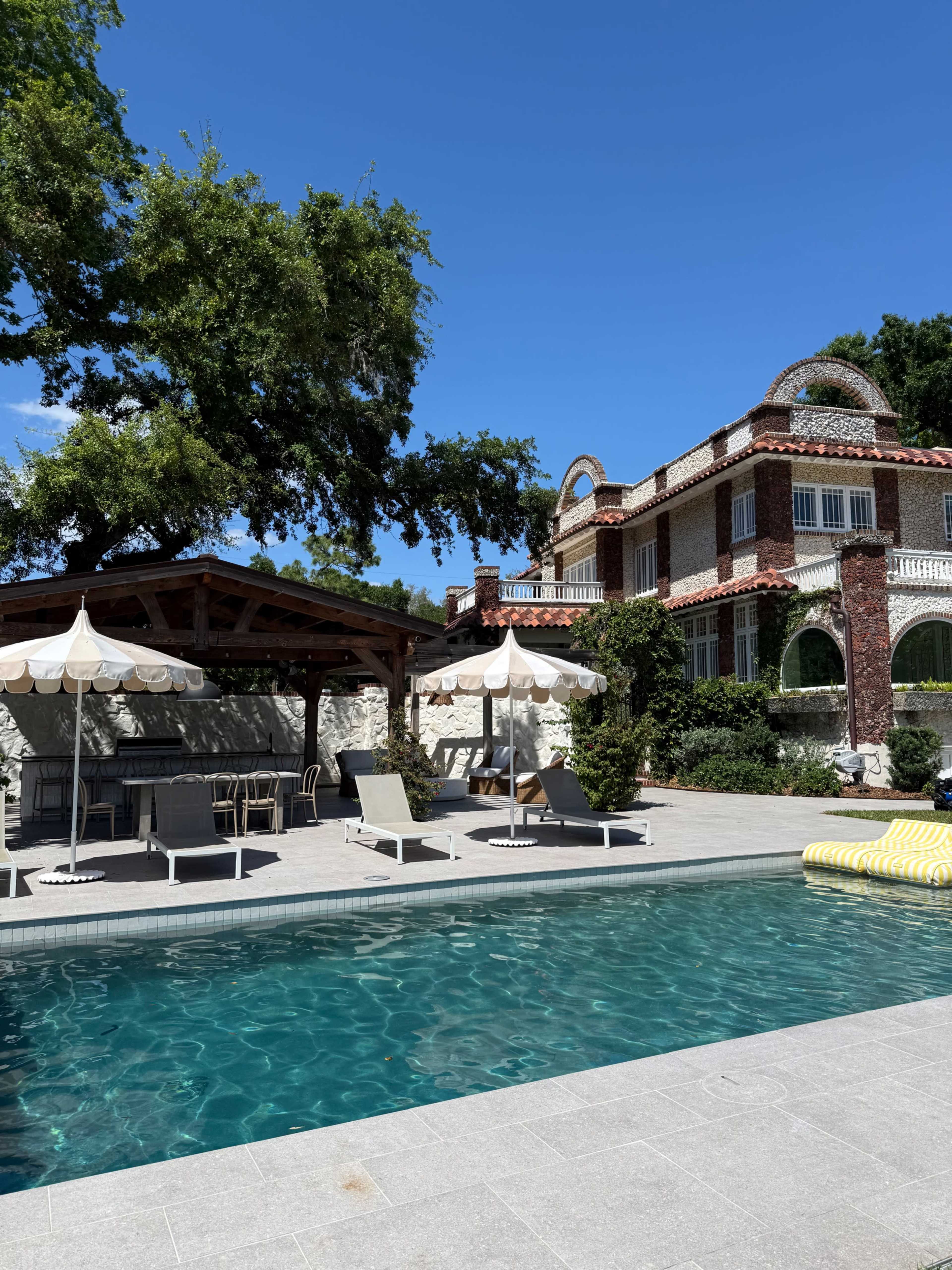 The image shows a luxurious pool area with lounge chairs, an outdoor dining space under a canopy, and a grand villa in the background surrounded by greenery.