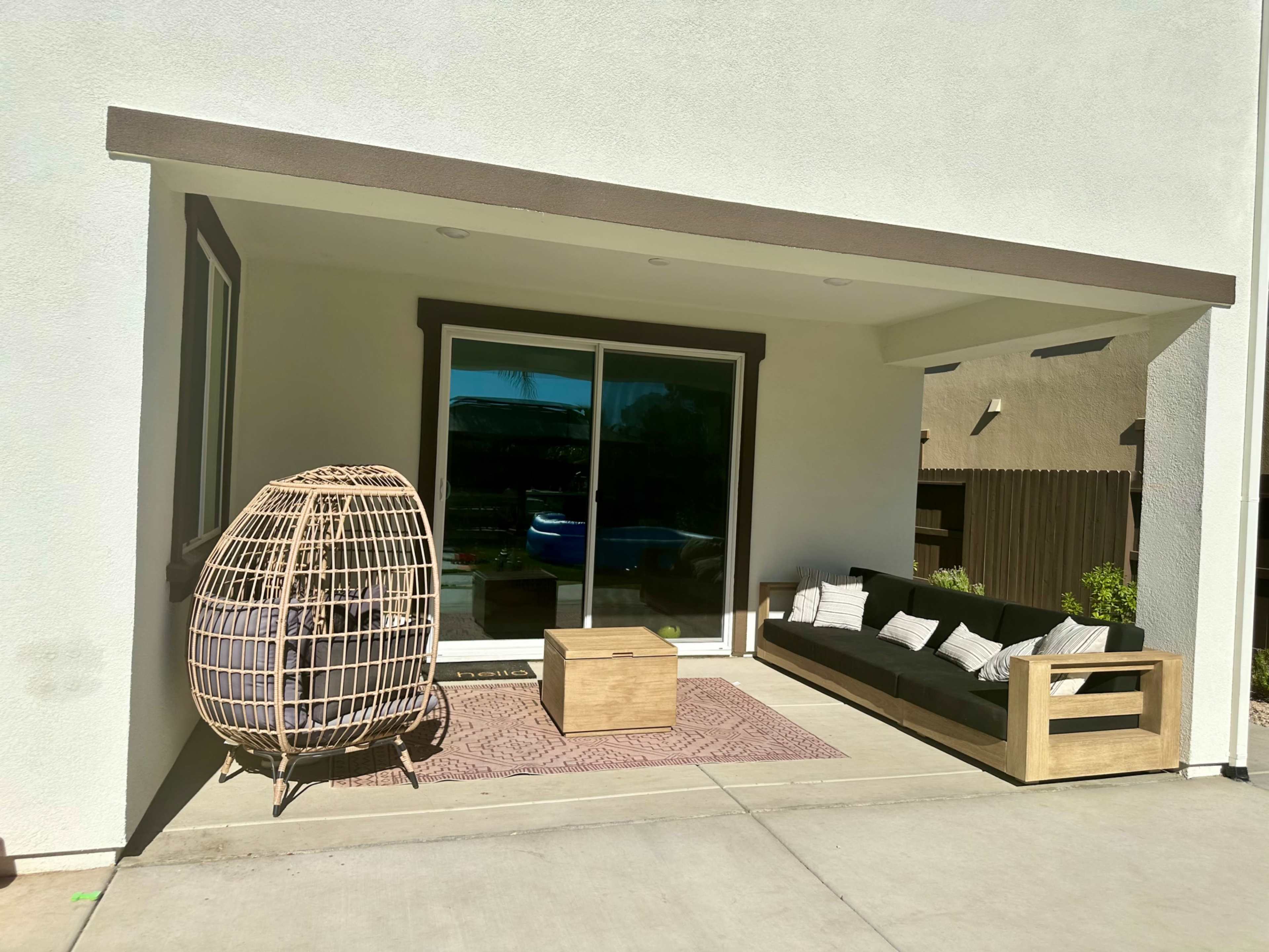 A modern patio features a wicker chair, a small wooden table, and a sectional sofa beneath a large overhang.