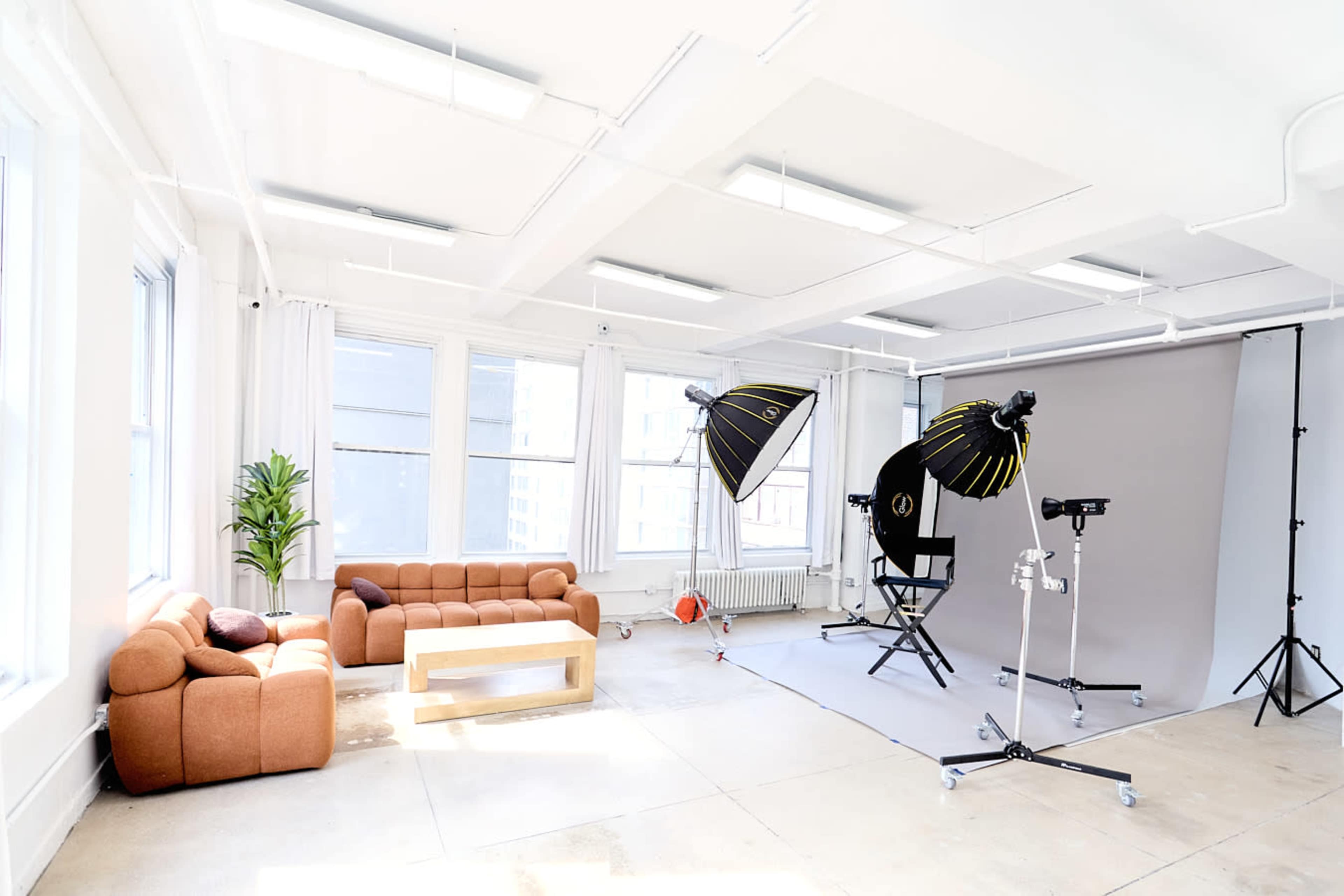 The image shows a bright photography studio featuring a brown sofa, a white coffee table, and two large lighting umbrellas positioned next to a gray backdrop.