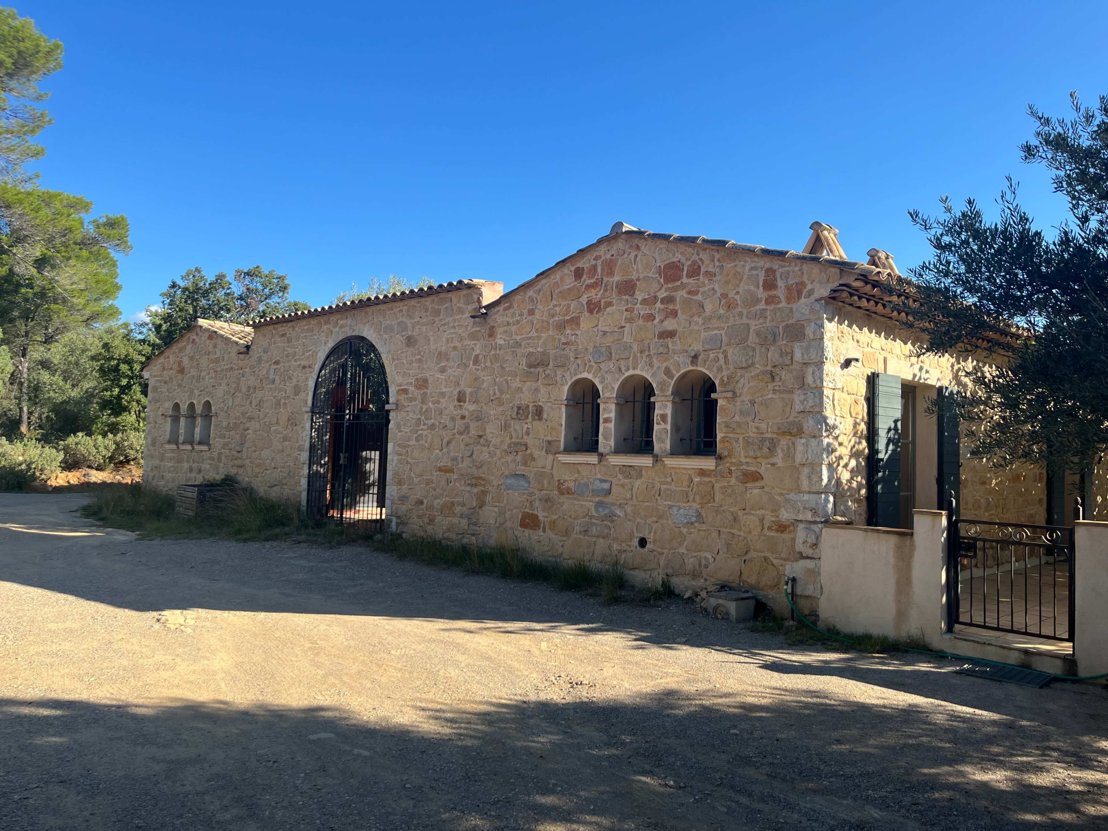 A stone building with arched windows and a gated entrance is set along a dirt road surrounded by trees.