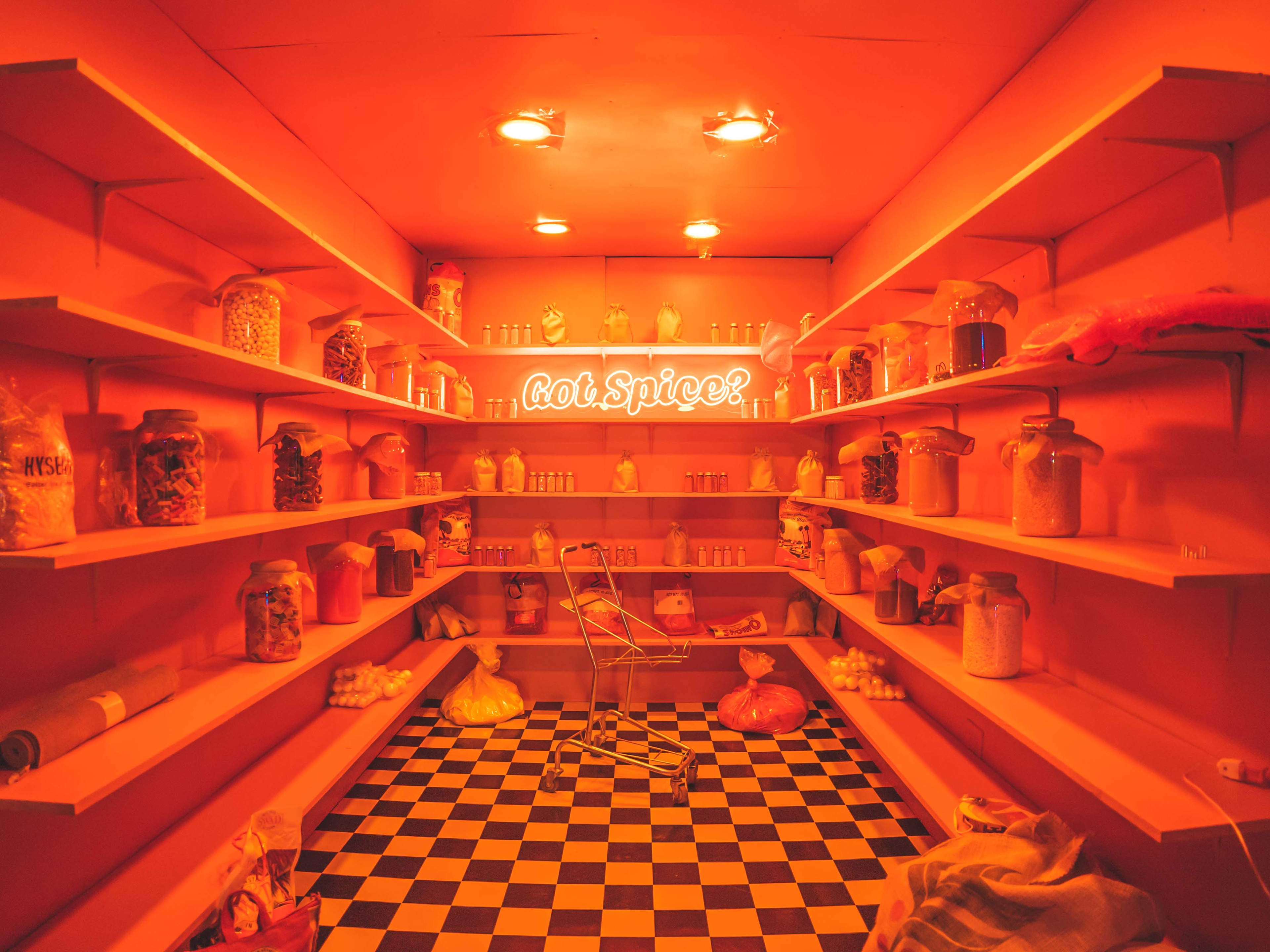 A brightly lit, orange-toned pantry features shelves lined with jars and containers of various ingredients, with a neon sign that reads "hot spice!" at the center.