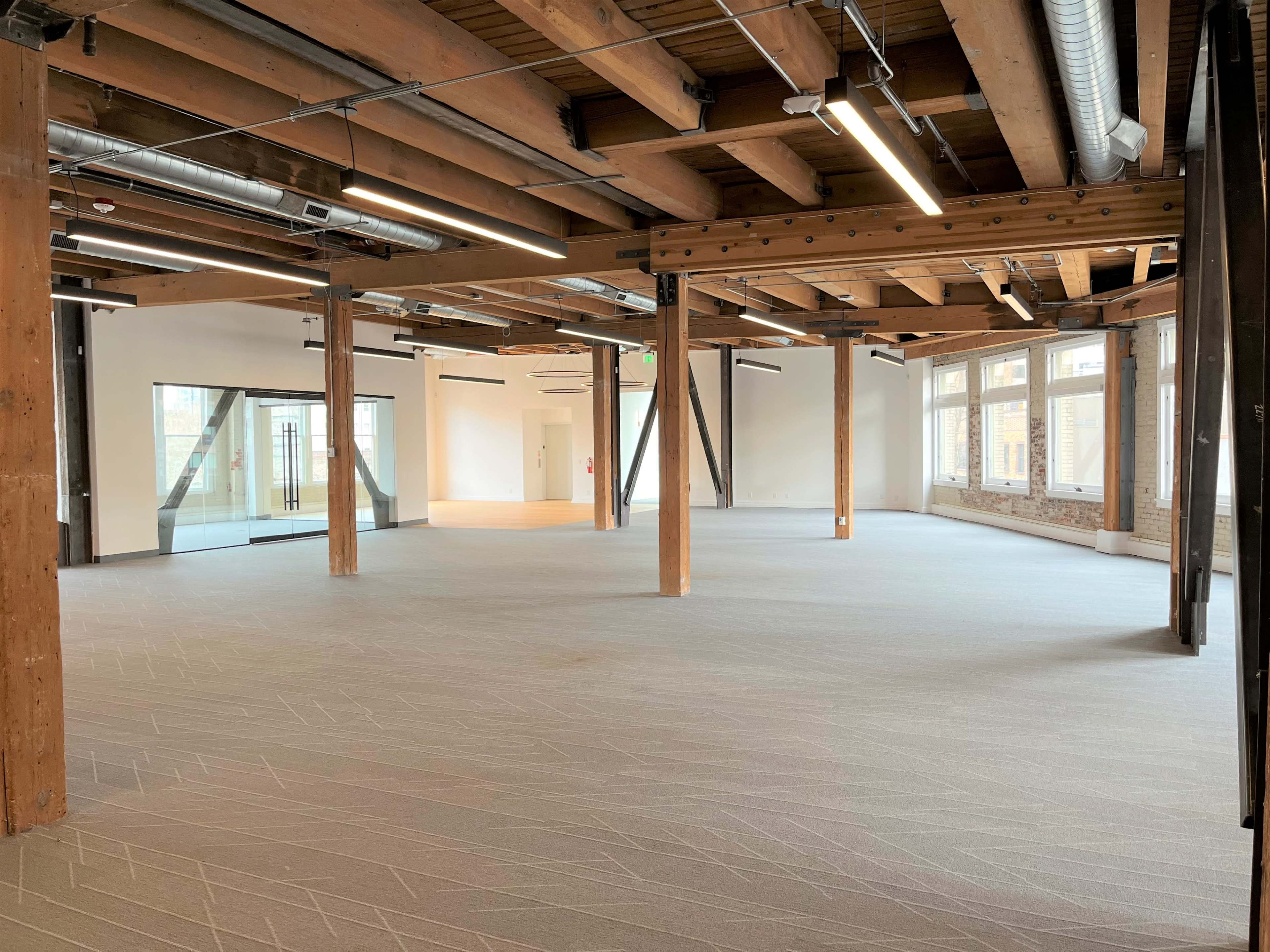 The image shows a spacious, empty room with wooden beams and large windows, featuring a light gray carpeted floor.