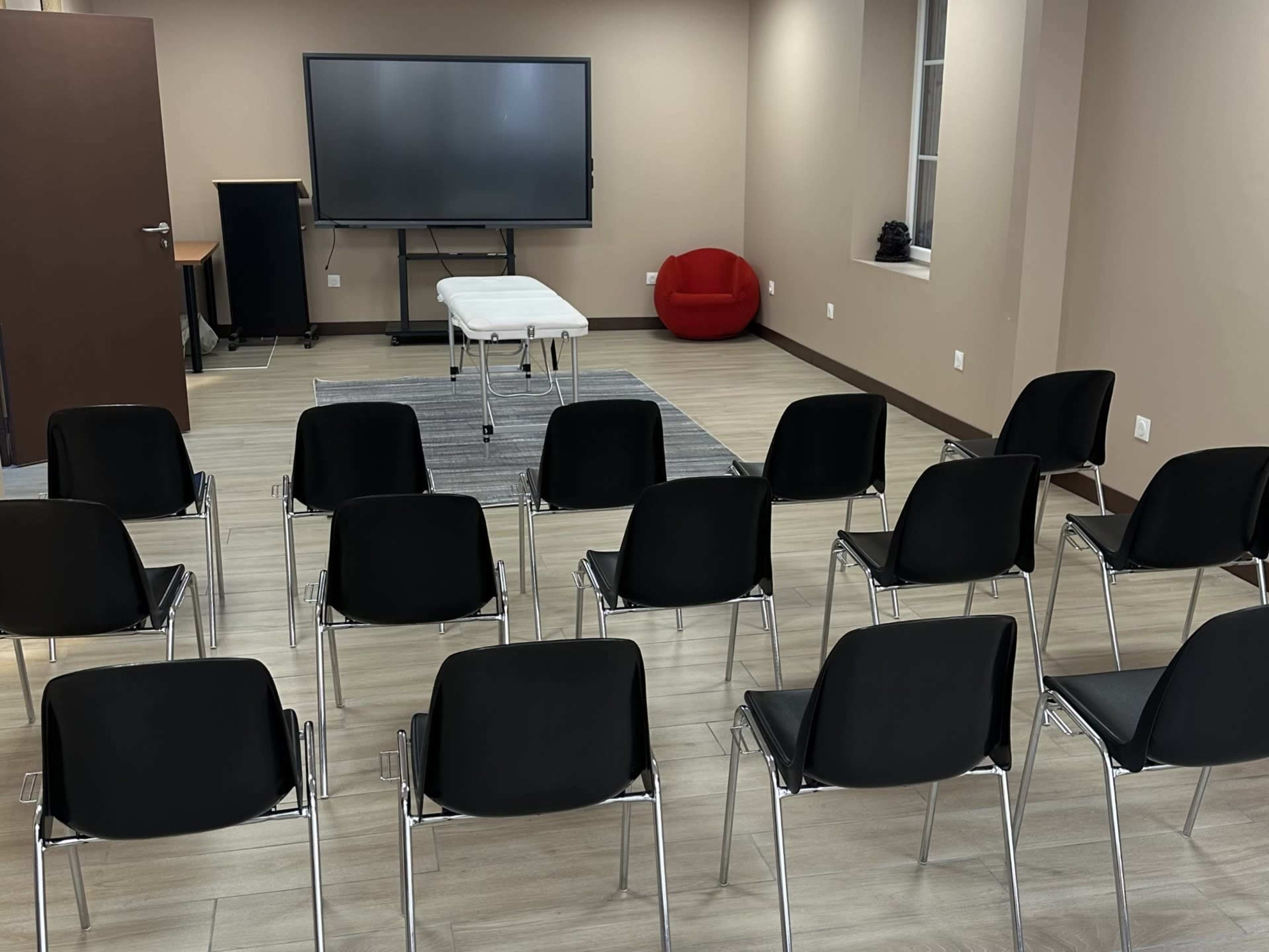The image shows a meeting room with several black chairs arranged in rows facing a table and a large screen at the front, with a red chair in the corner.