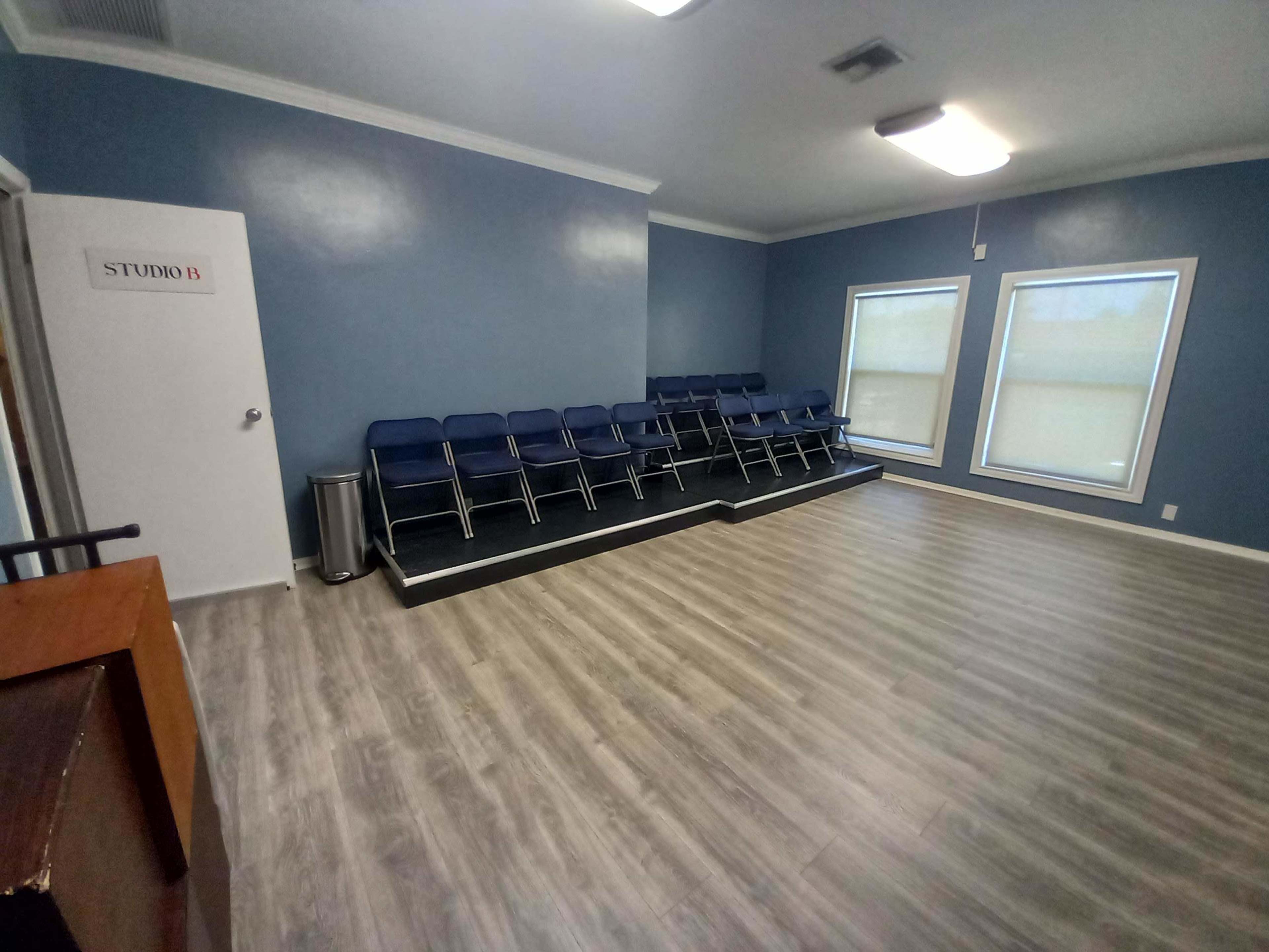 A waiting room with a row of blue chairs against a wall and two large windows, all on a wooden floor.