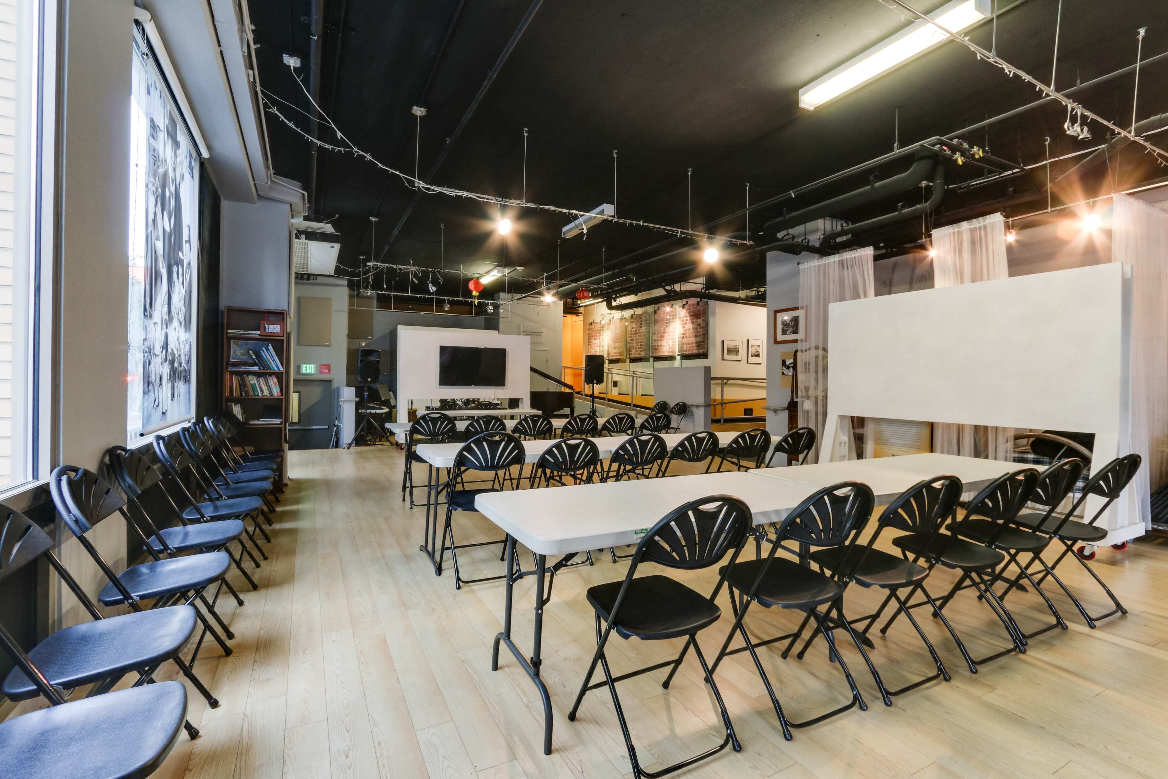 The image shows a spacious room set up with several rows of folding chairs facing a series of tables, with a projector screen and artworks displayed on the walls.