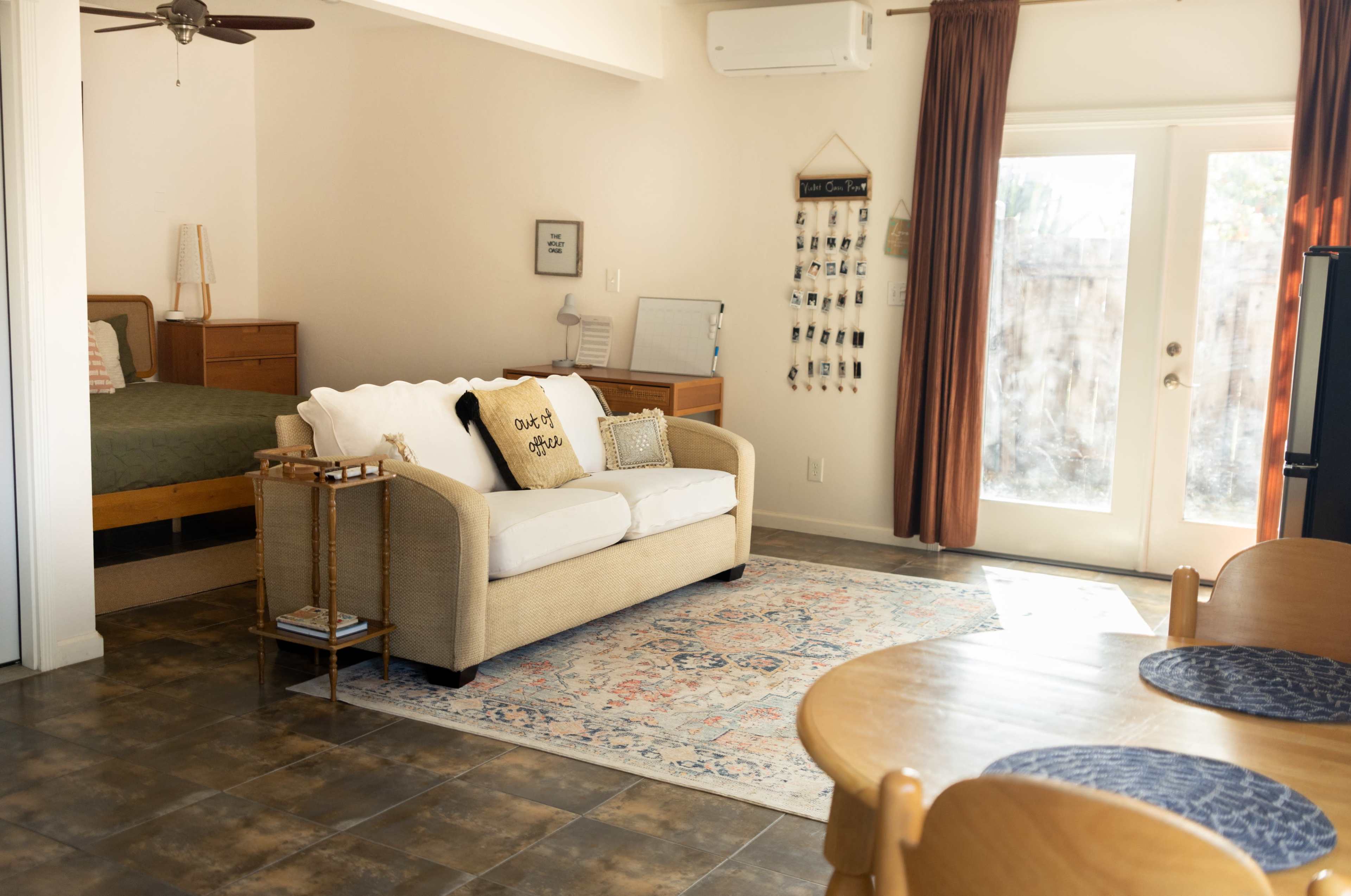 A living space features a sofa facing a small table, with a bed in the background and large windows providing natural light.