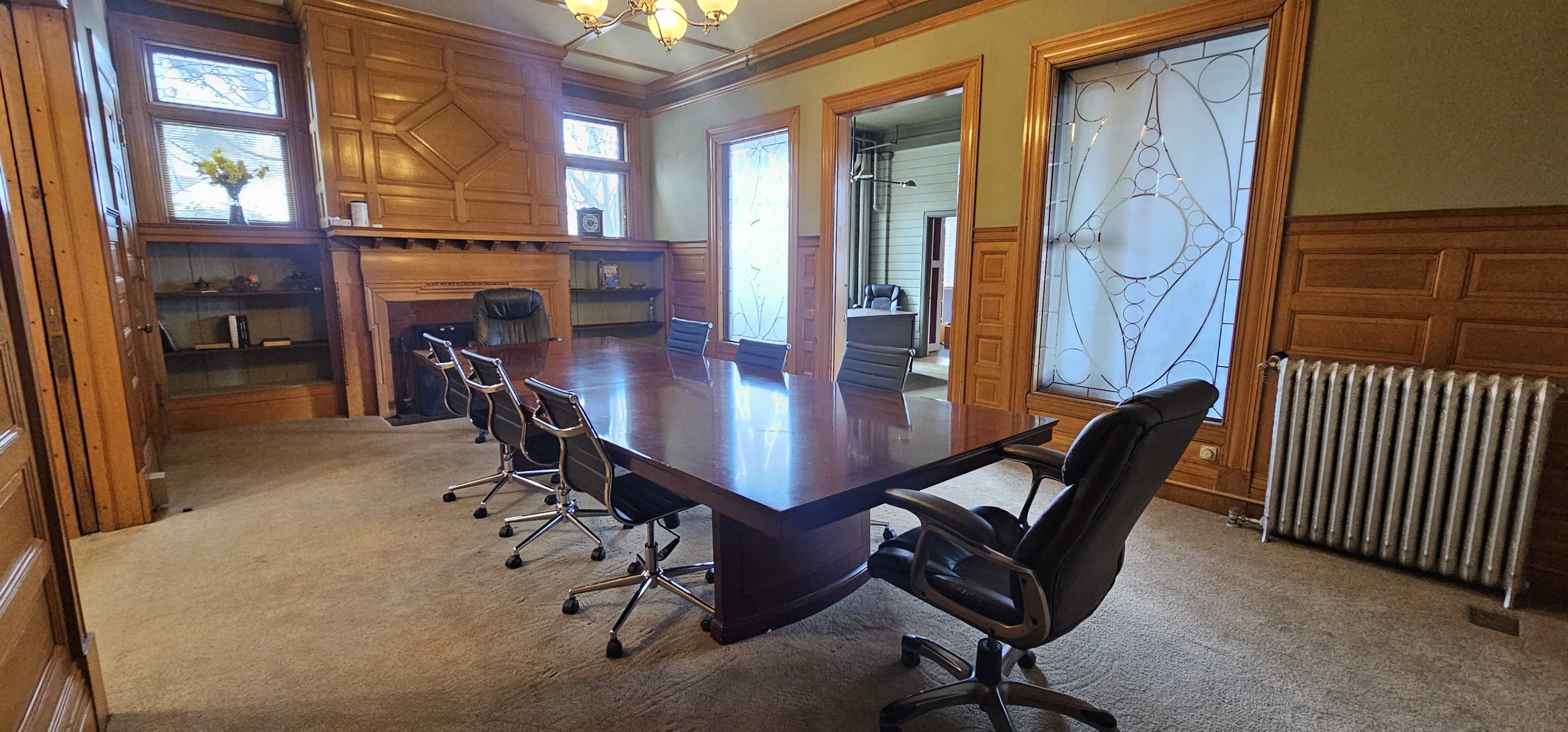 A large conference room with a wooden table, six rolling chairs, and a decorative fireplace, framed by windows and wooden paneling.