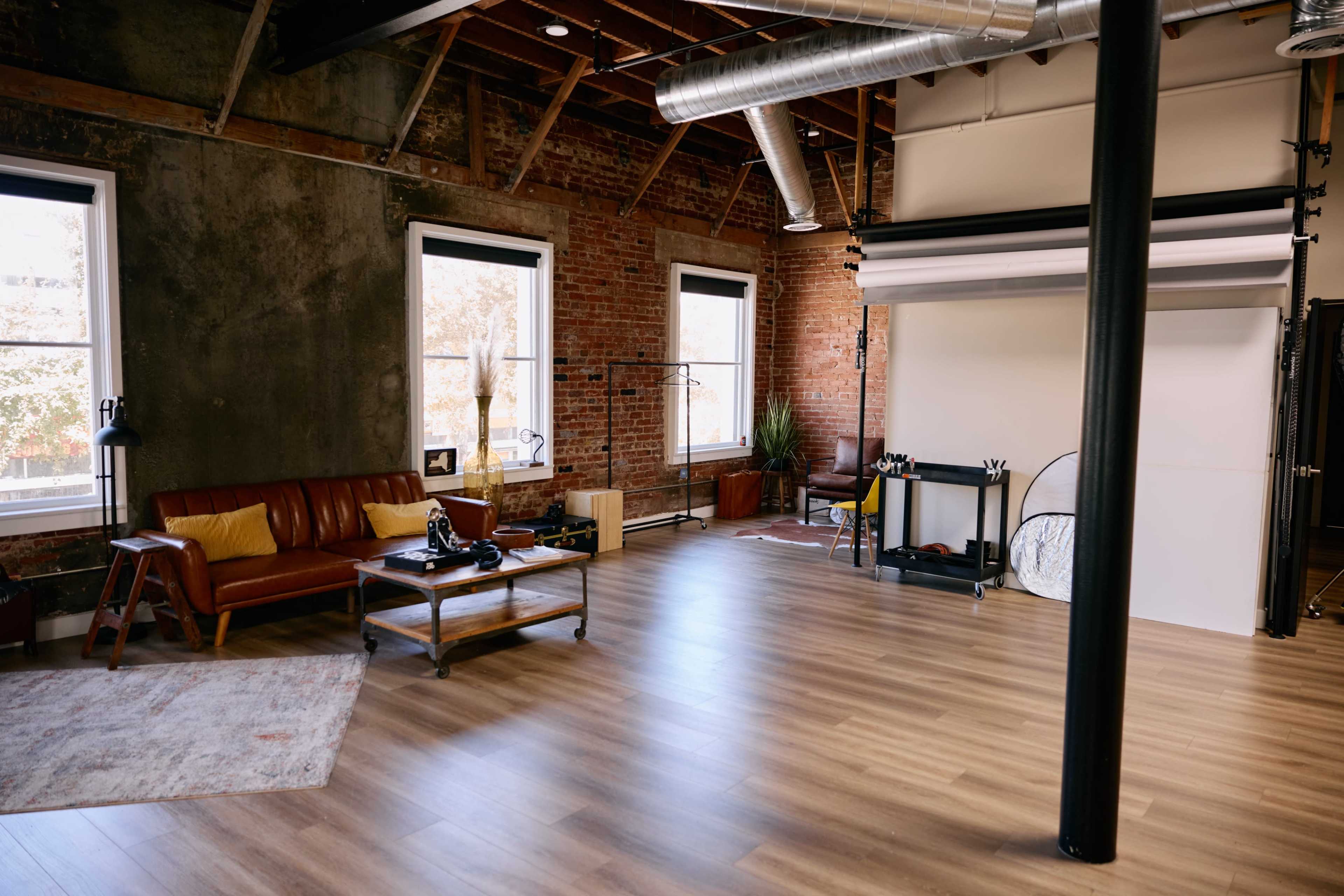The image shows a spacious studio with hardwood flooring, brick walls, a leather sofa, and a photography backdrop setup.