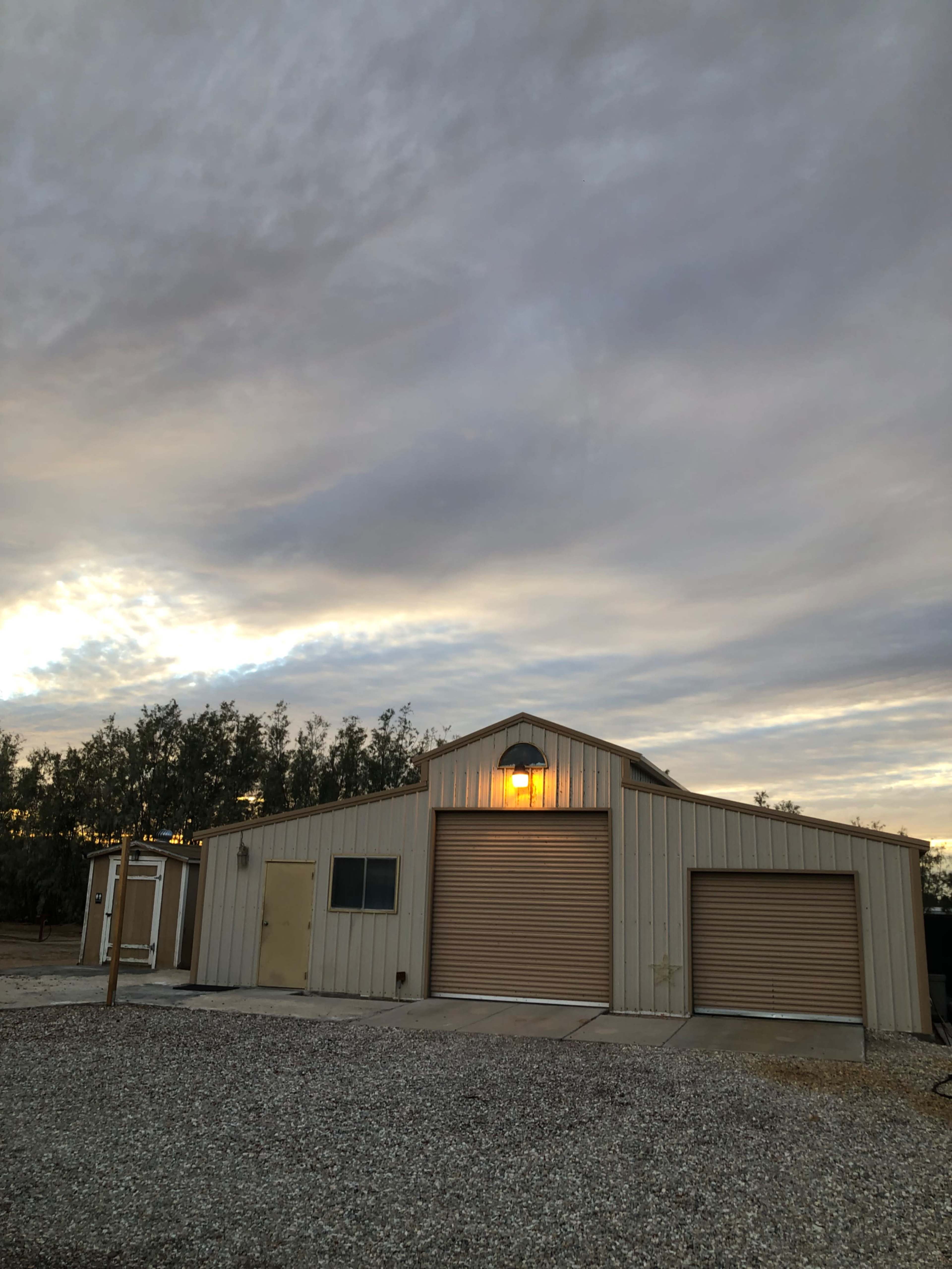A metal building with two garage doors and a lit exterior light stands on a gravel surface under a cloudy sky.