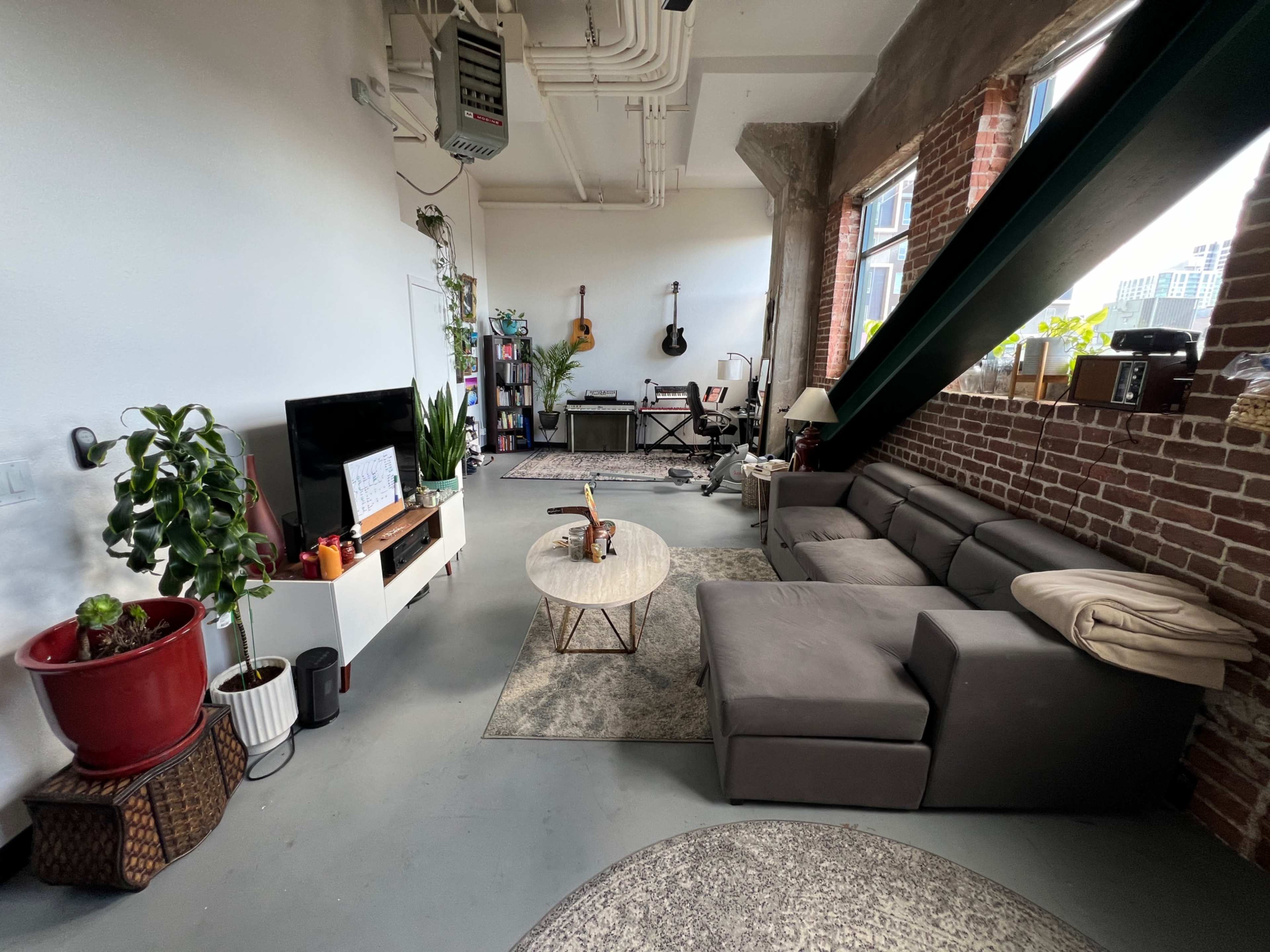 The image shows a modern living room with a sectional couch, a coffee table, a television, potted plants, and musical instruments near a window with exposed brick walls.