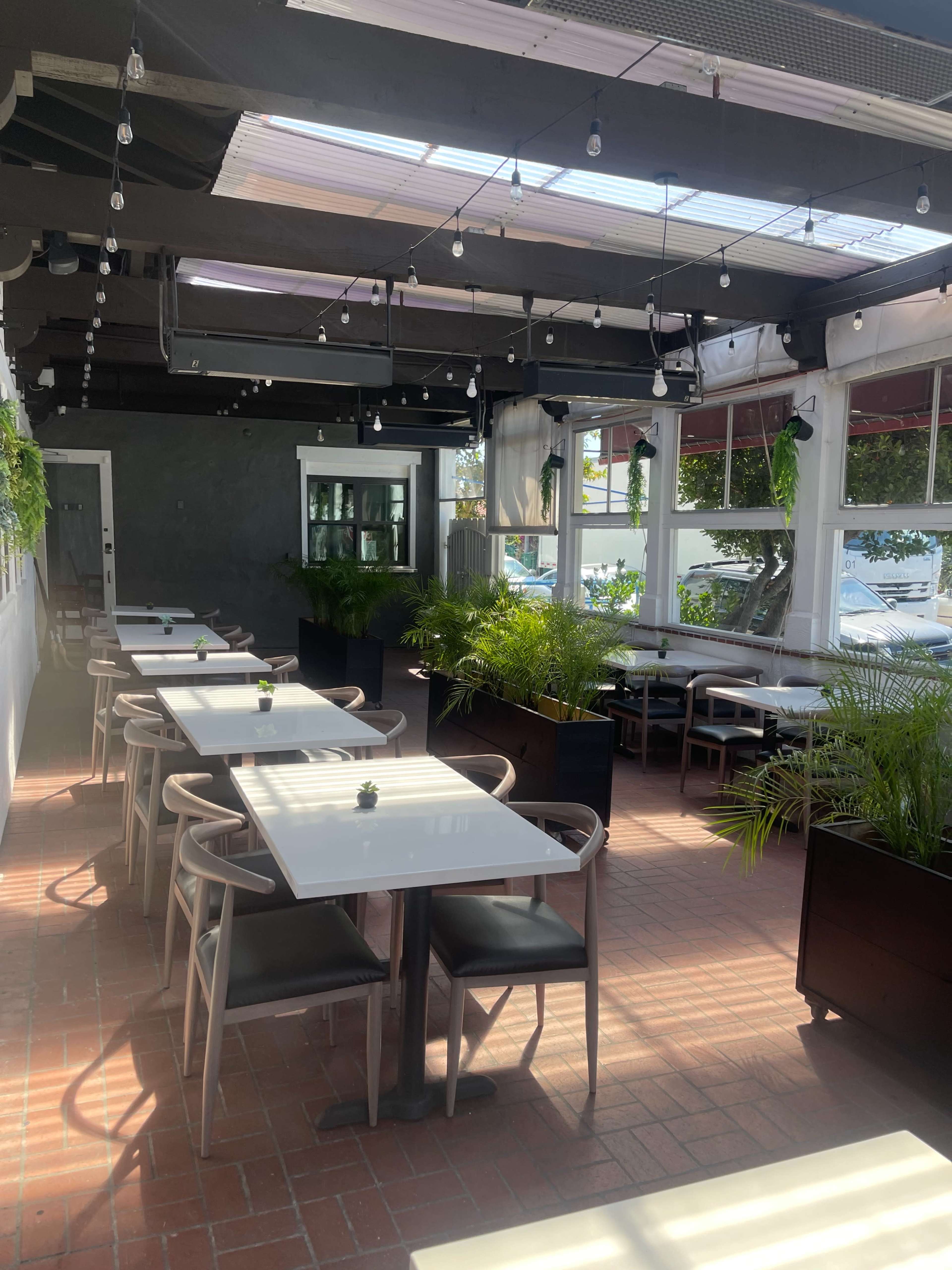 A bright indoor dining space featuring white tables, black chairs, and greenery arranged around large windows.