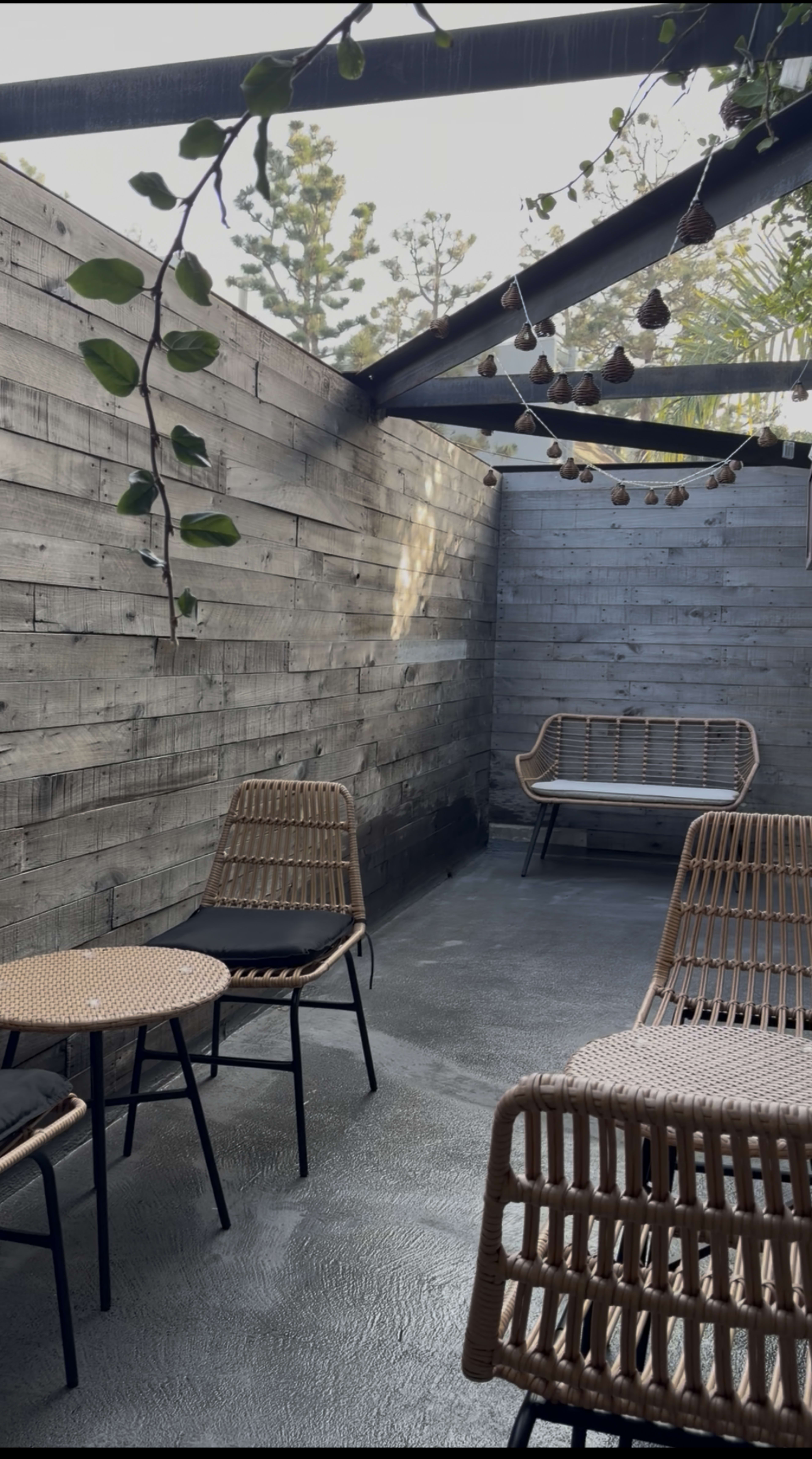 A small, minimalist outdoor seating area features wicker chairs and a low table, surrounded by wooden walls and a roof of open beams.