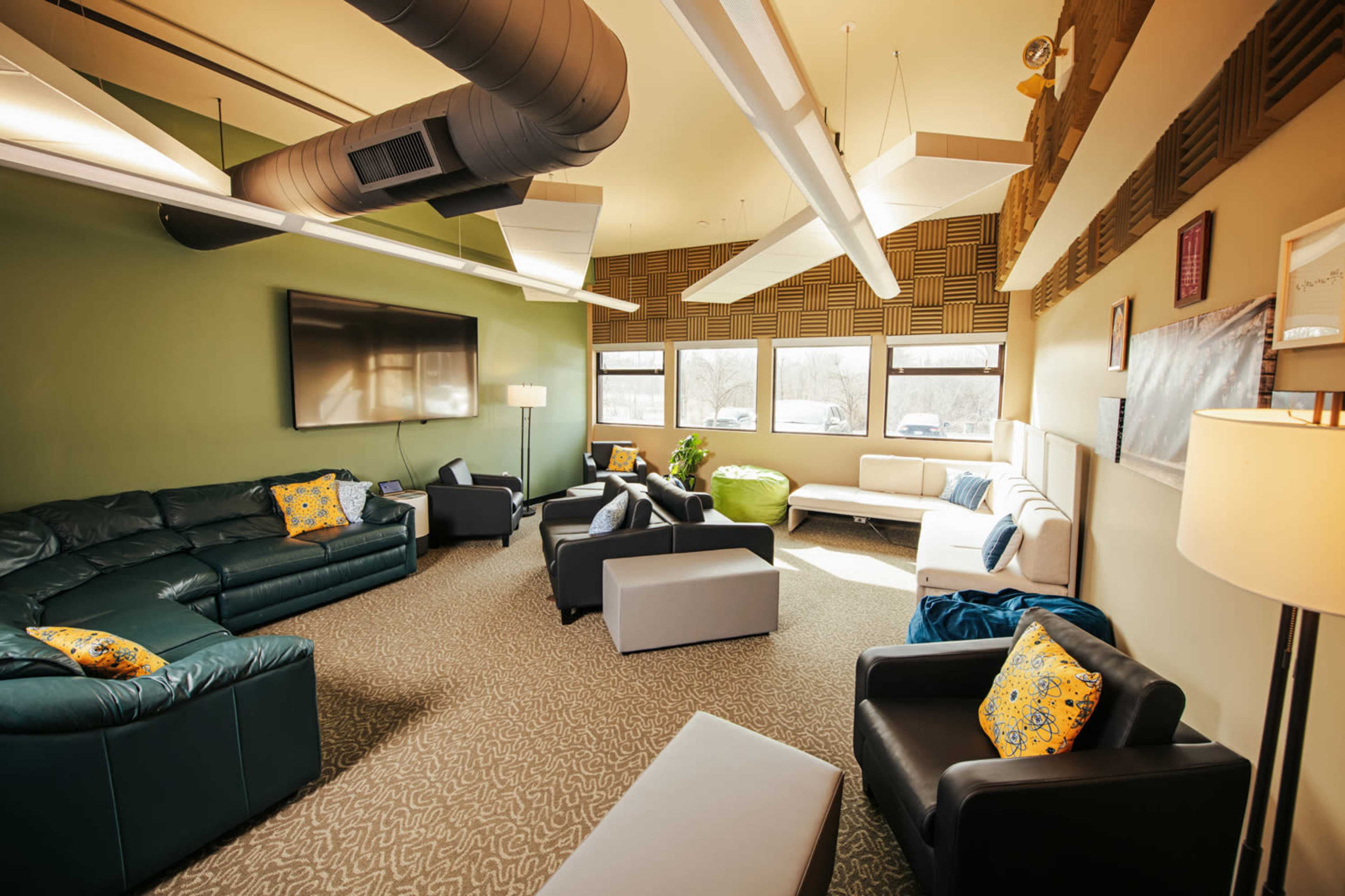 The image shows a modern lounge area with various seating options, including sofas and armchairs, alongside a large television mounted on the wall.