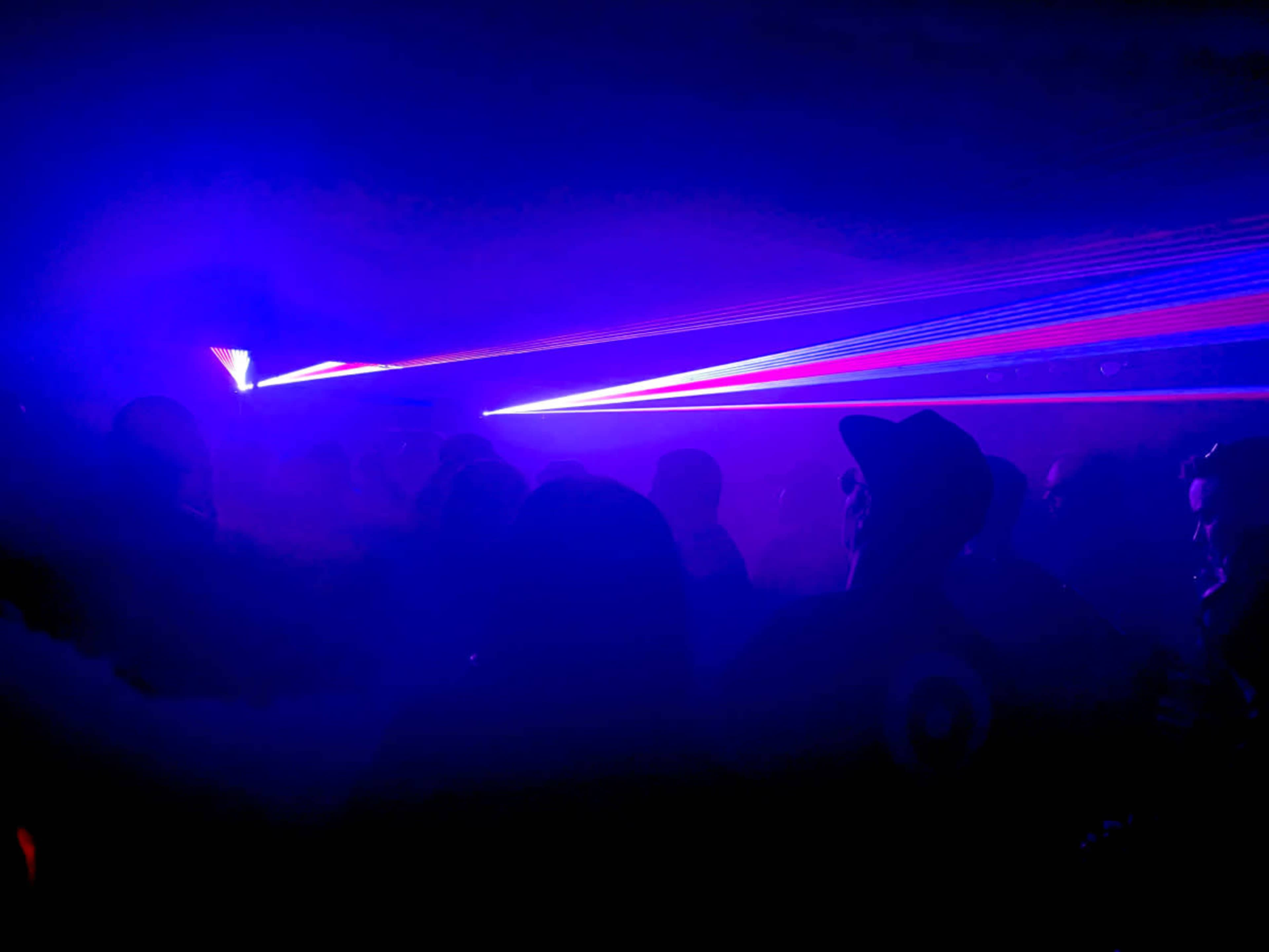 A dimly lit space filled with silhouetted figures, illuminated by vibrant purple and blue laser beams cutting through a haze of smoke.