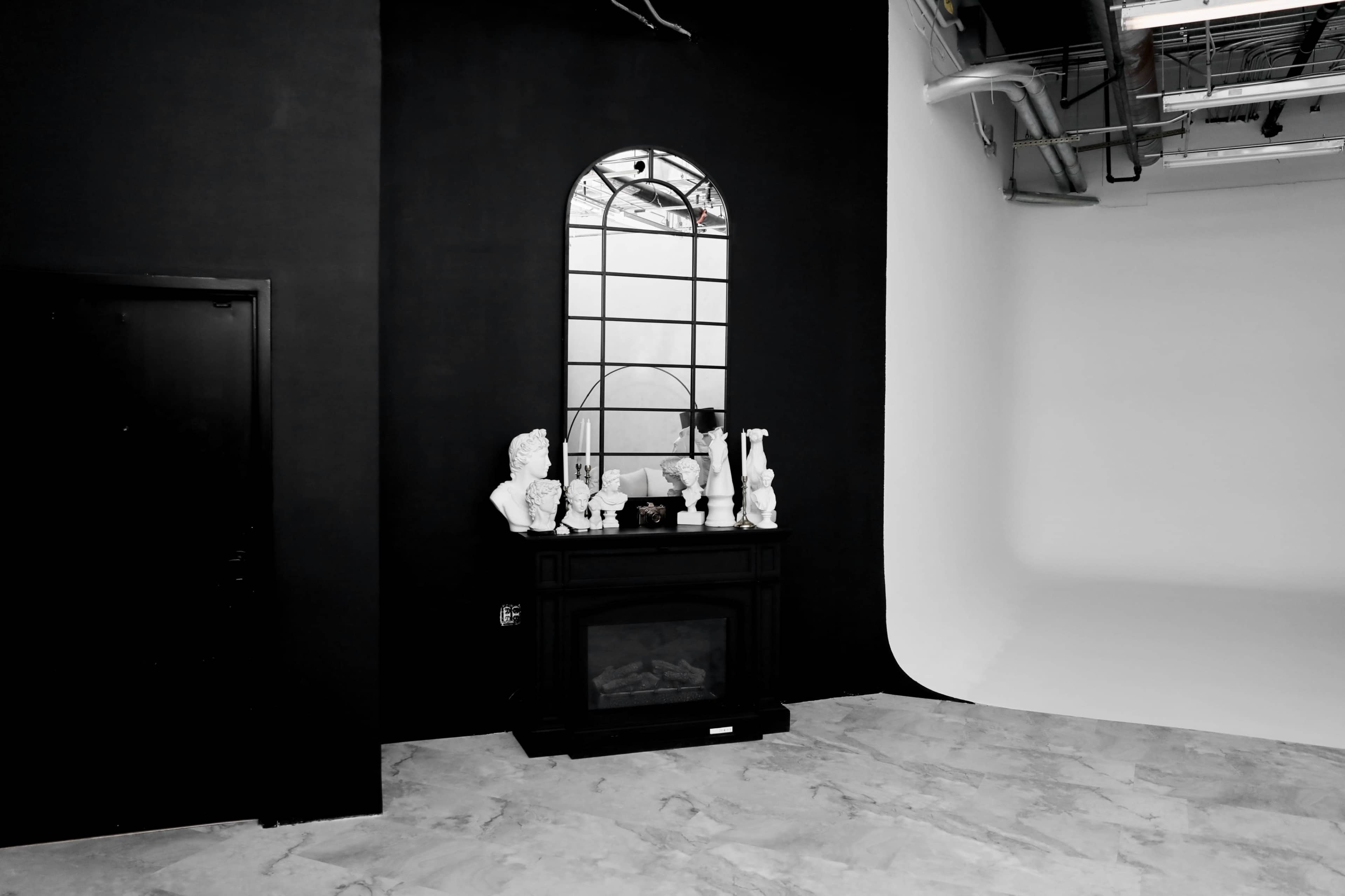 A minimalist studio space featuring a black wall, a decorative mirror above a dark wooden mantelpiece with white sculptures, and a contrasting white backdrop on the opposite side.