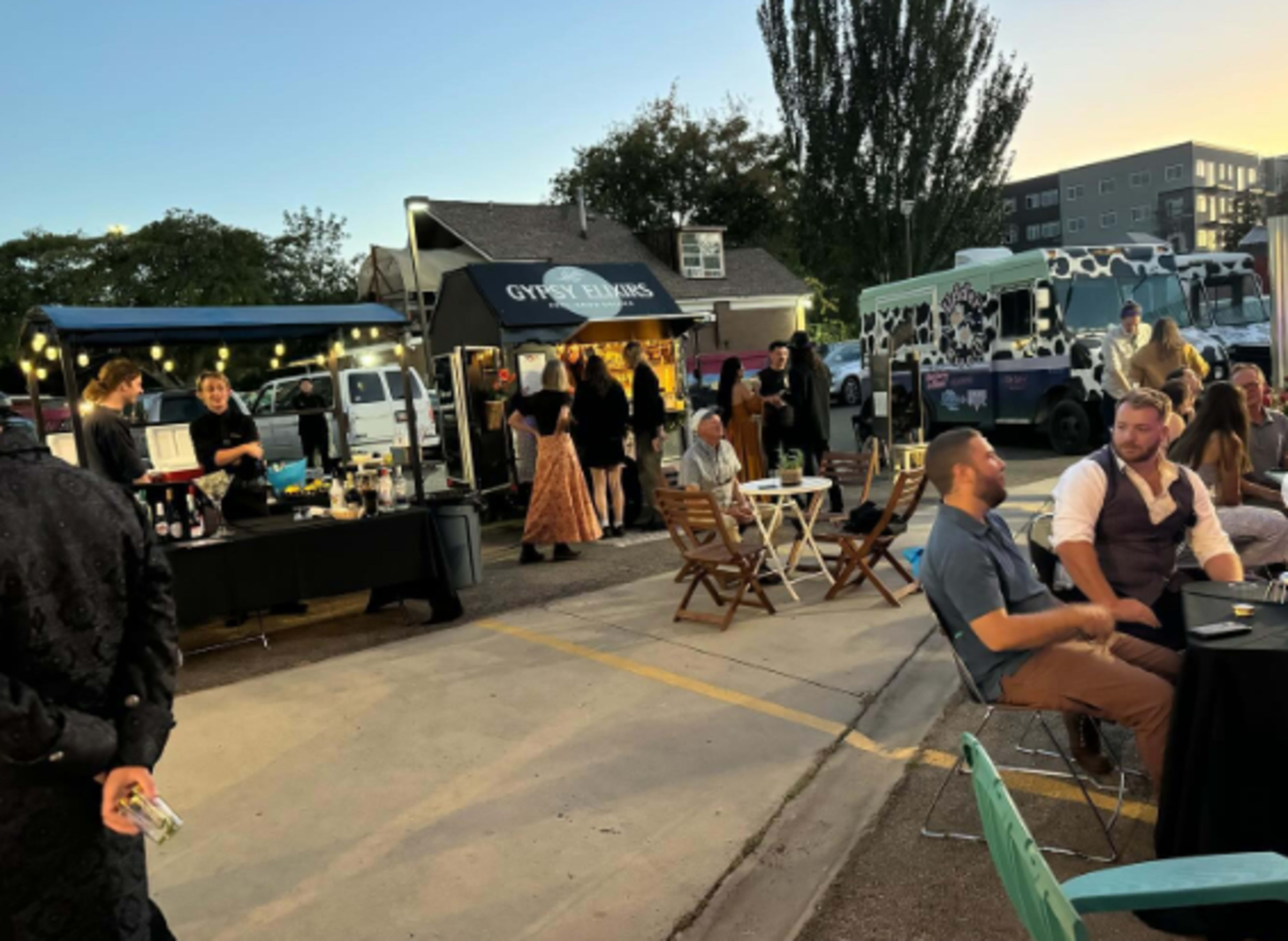A lively outdoor event features food trucks and guests socializing at tables as night falls.