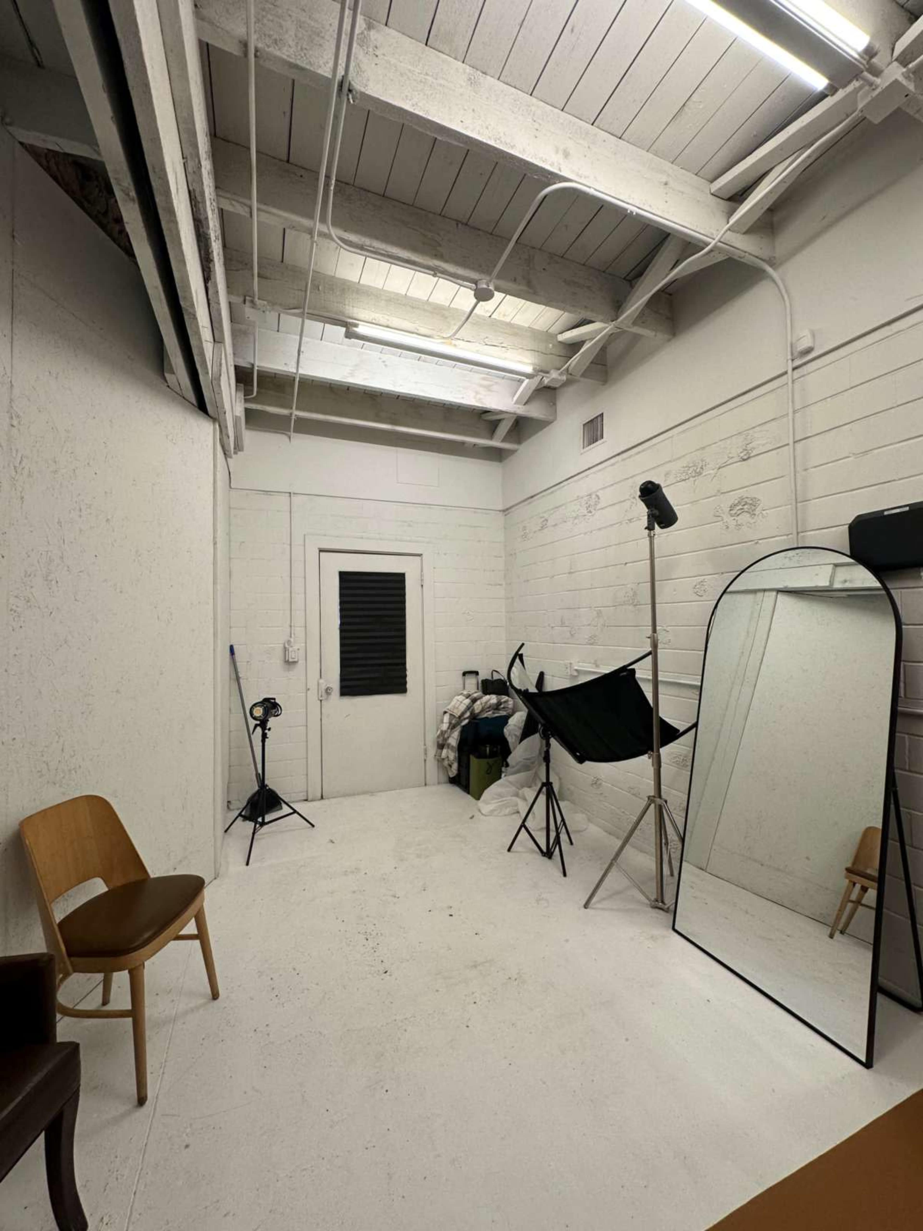 The image shows a sparsely furnished studio space with white walls, a hardwood chair, a large mirror, and photography equipment including a light stand and an umbrella reflector.