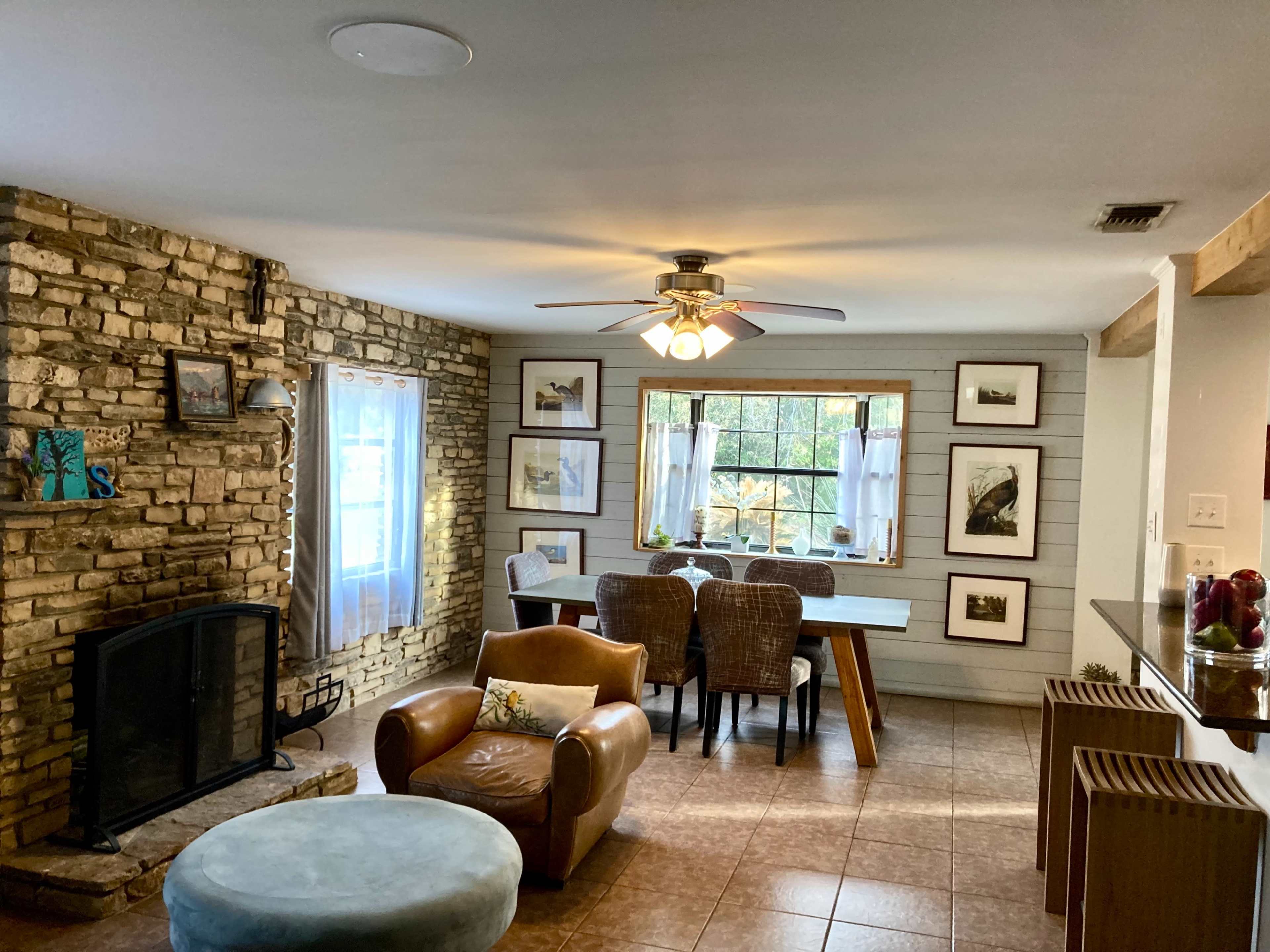 A cozy living space featuring a stone fireplace, a dining table with wicker chairs, and large windows with white curtains.