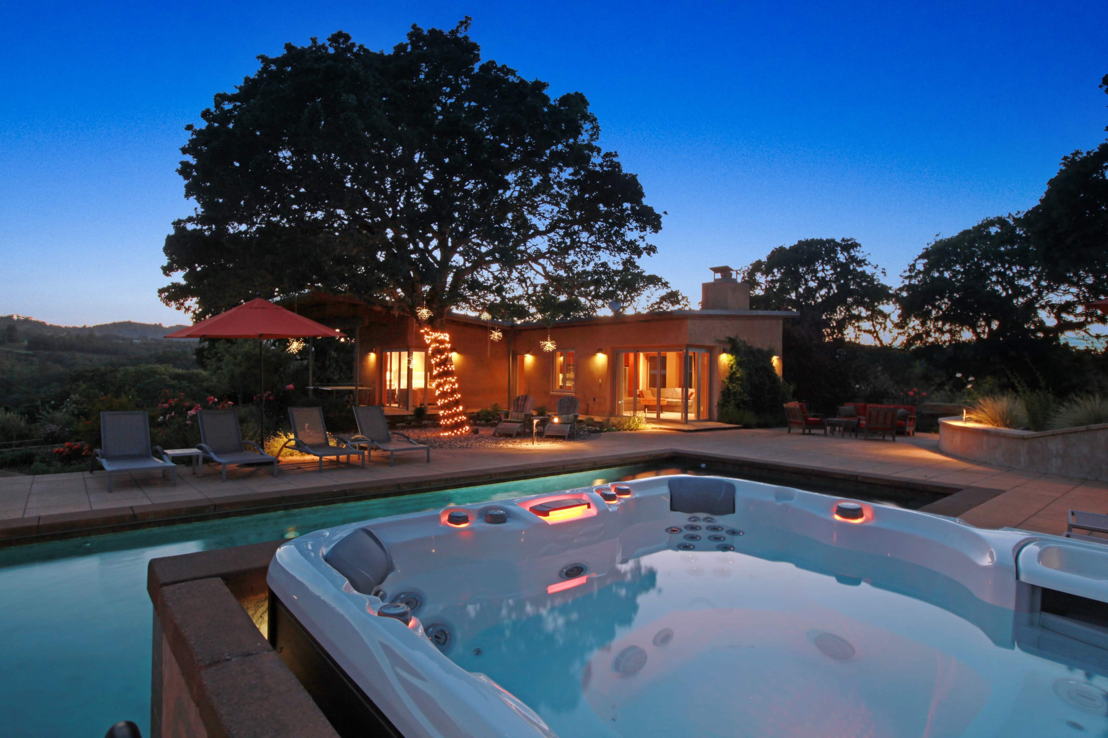 A hot tub beside a swimming pool, with a house and trees illuminated in the evening sky.