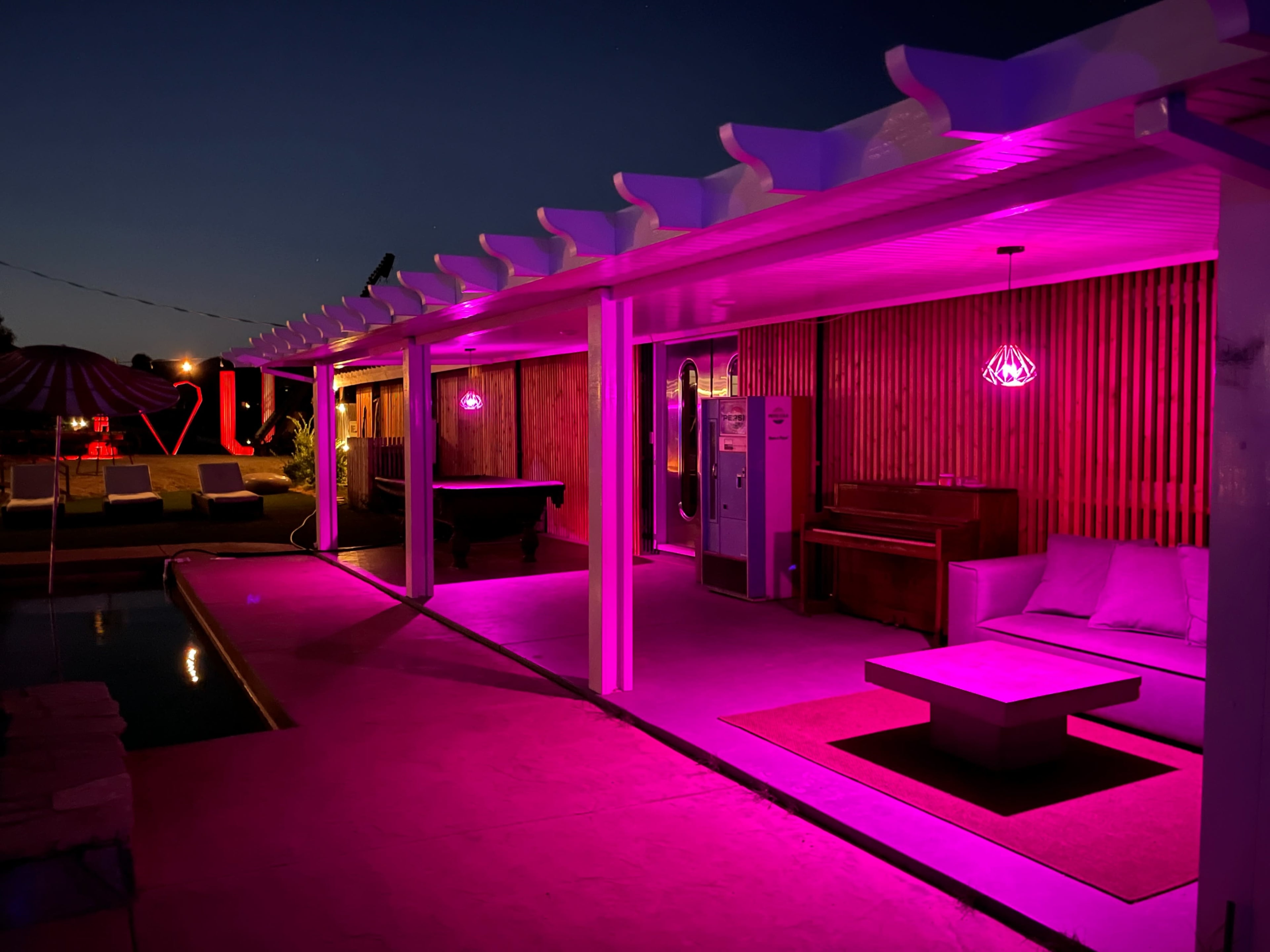 The scene features a modern outdoor lounge area illuminated by pink lights, with a pool, seating, and a piano under a covered structure.