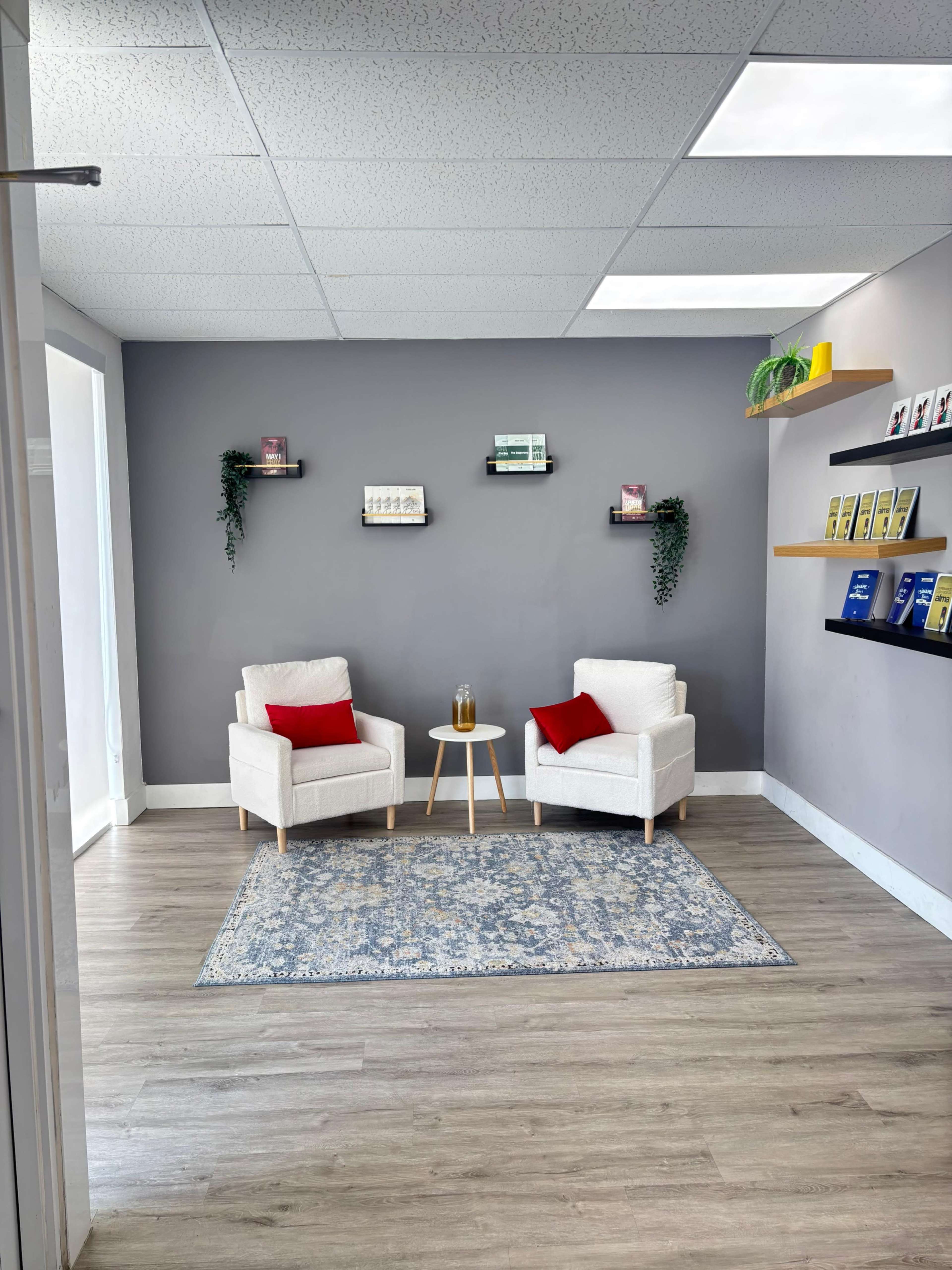 The image shows a modern waiting area with two white armchairs, a small wooden side table, and decorative wall shelves displaying books and plants against a gray wall.