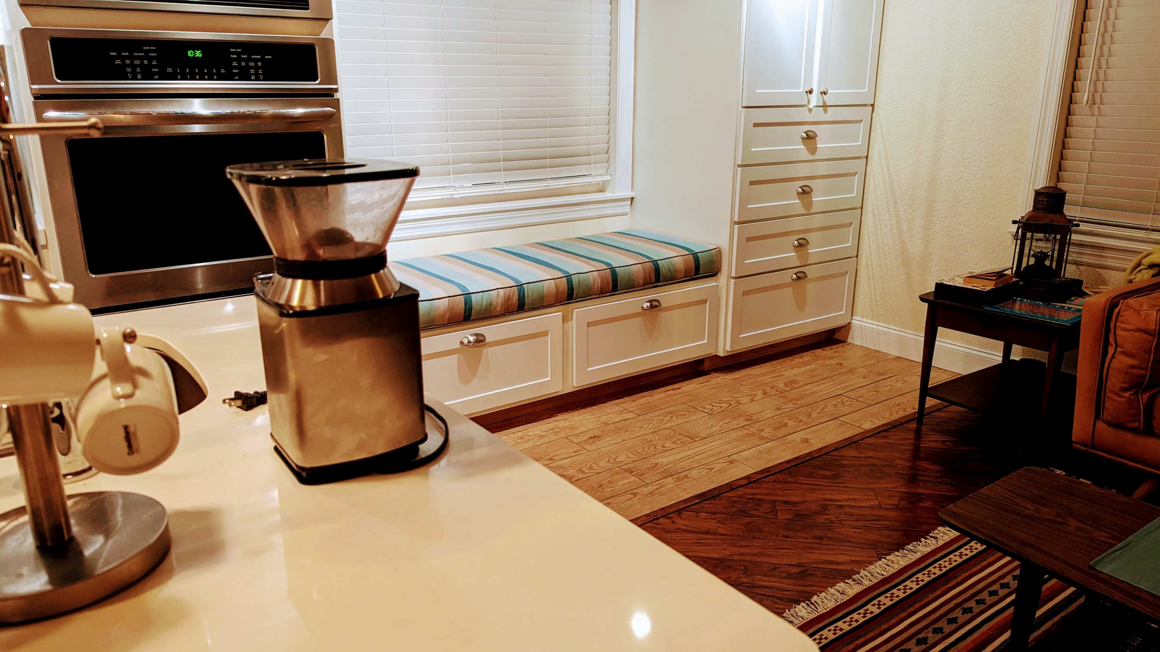 A kitchen scene features a countertop with a coffee grinder, an oven, and a seating area with a striped cushion beside storage cabinets.