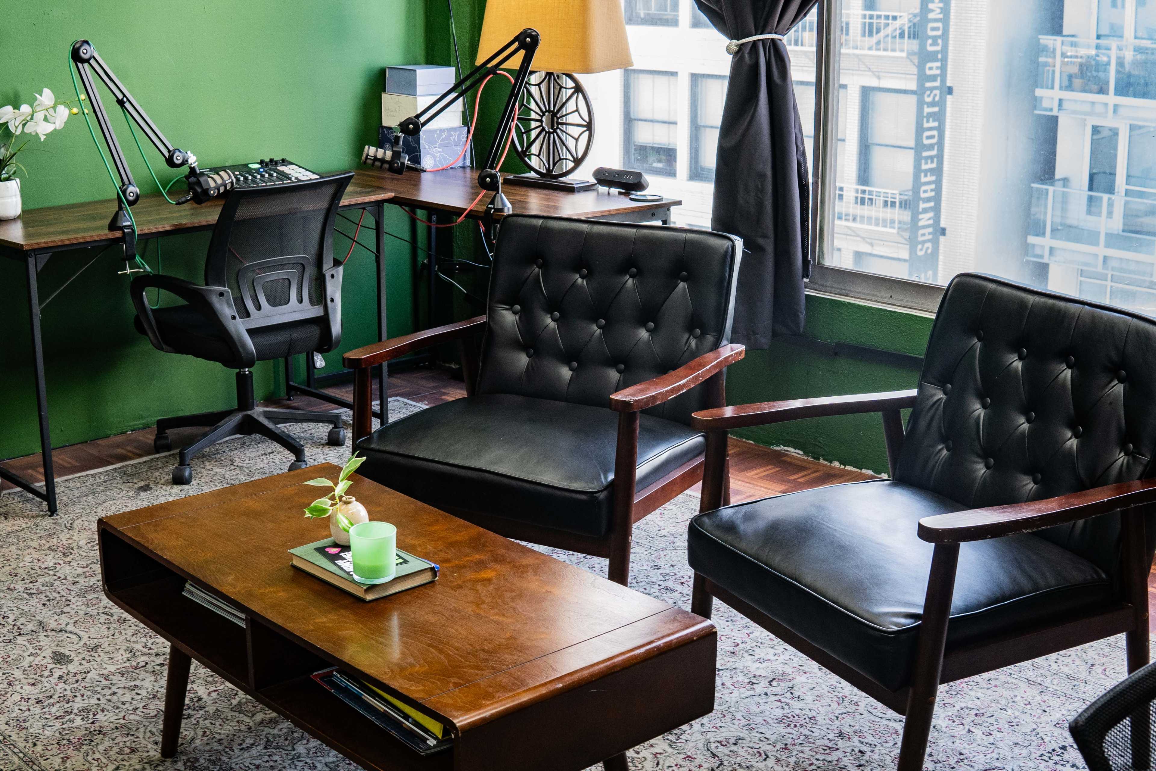 The image shows a modern workspace with two black chairs, a wooden coffee table, and a desk equipped with a microphone and recording equipment, set against a green wall with large windows.