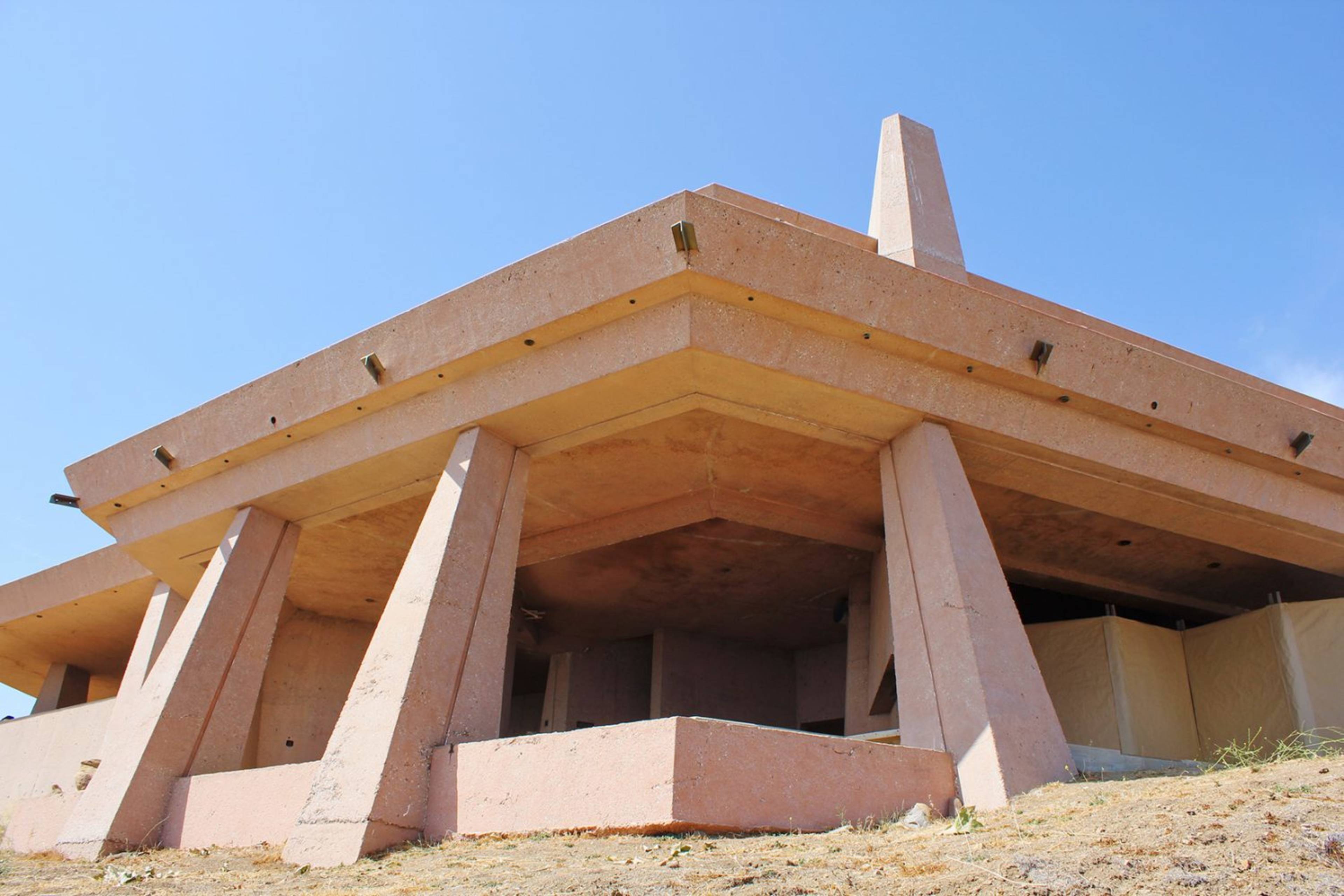 The image shows a large, unfinished building with a flat roof supported by tall, angular columns against a clear blue sky.