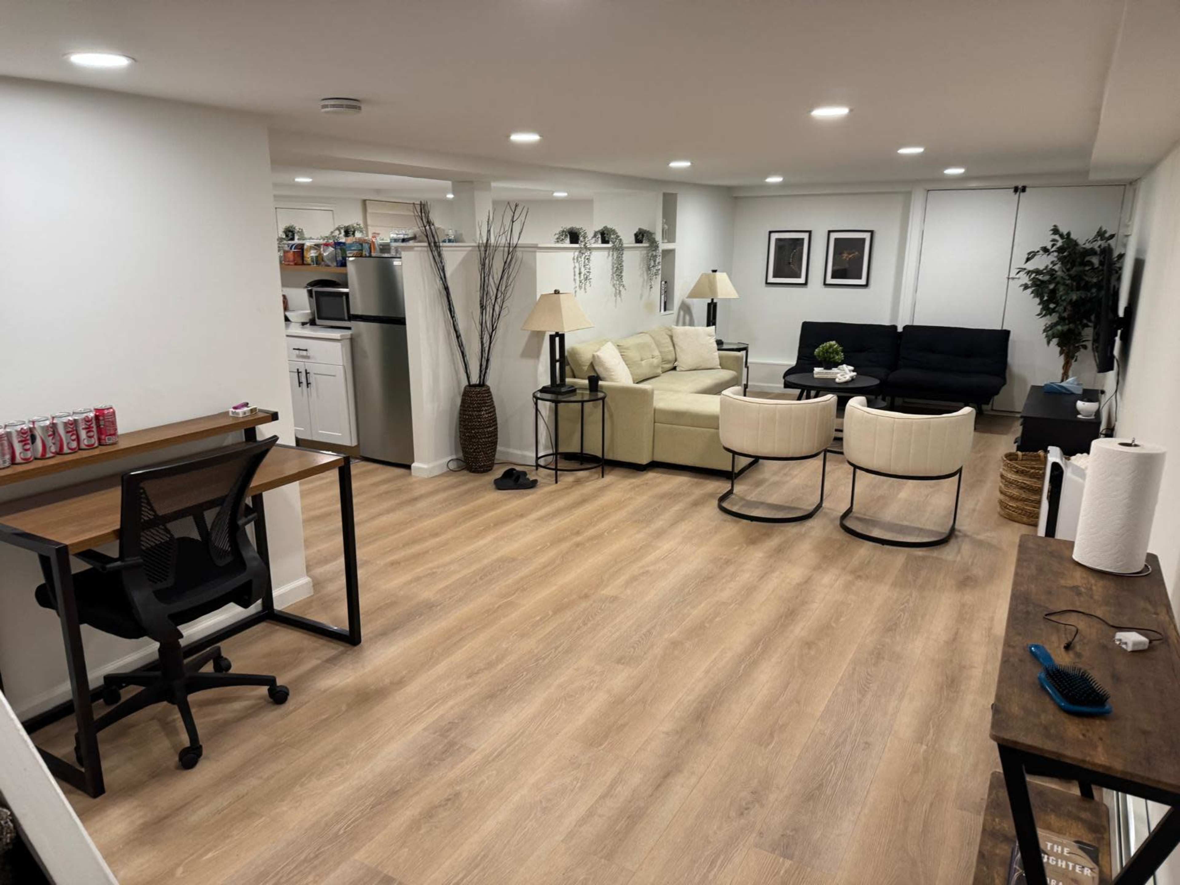 The image shows a well-lit basement living area featuring a sofa, armchairs, a desk, a small kitchenette, and decorative plants.