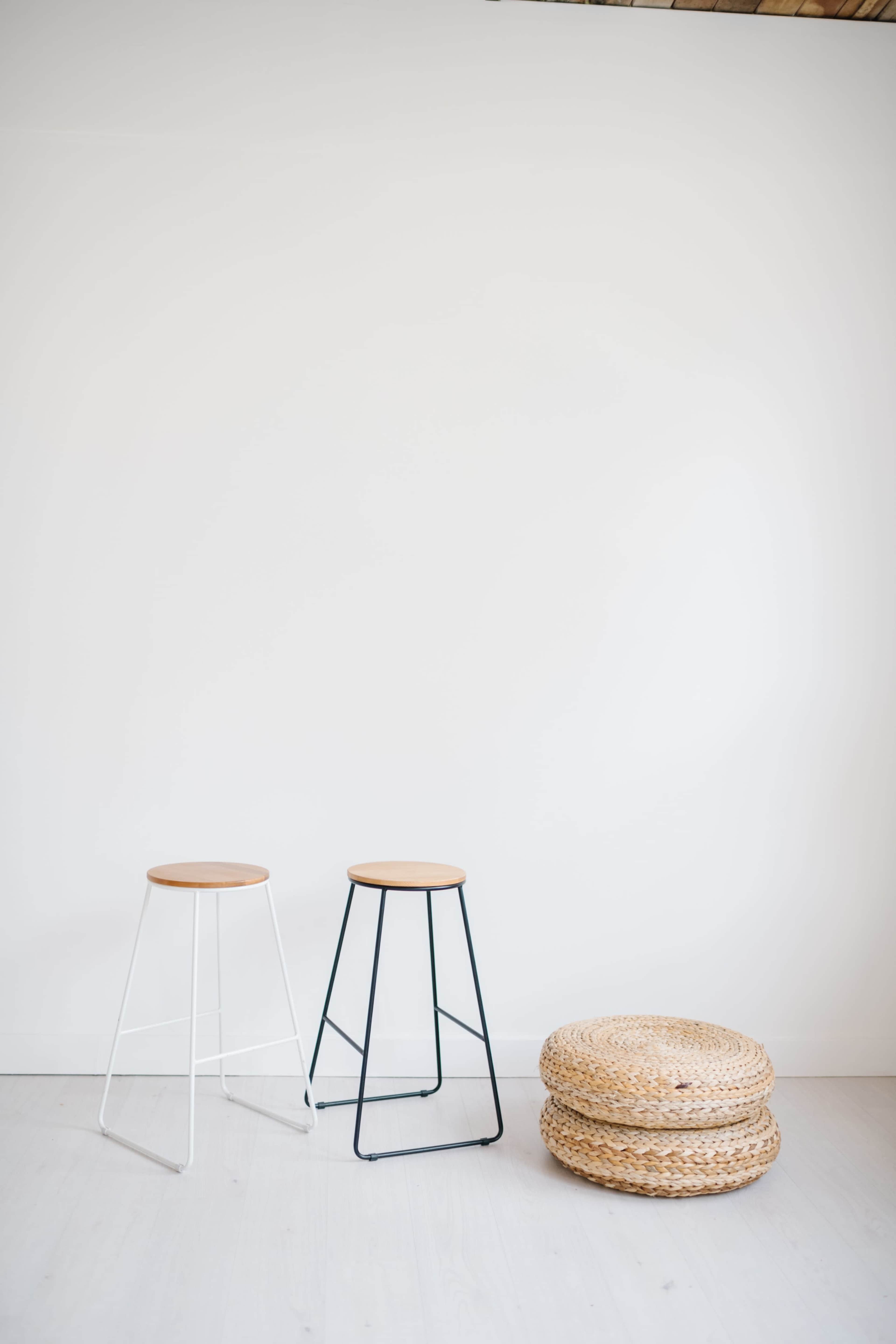 Two stools with wooden tops and metal frames, one white and one black, are positioned beside three woven baskets on a light wooden floor against a plain white wall.