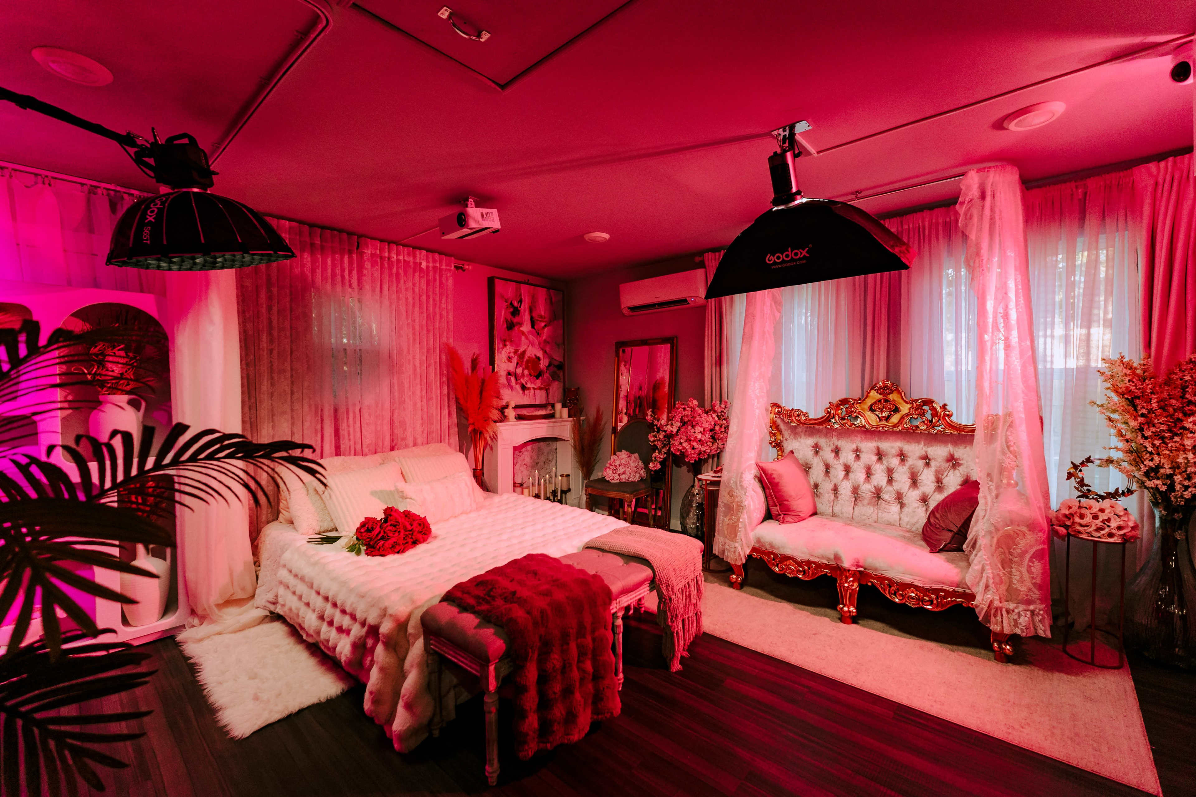 The image shows a cozy room with a bed and an ornate couch, decorated with pink lighting and floral arrangements.