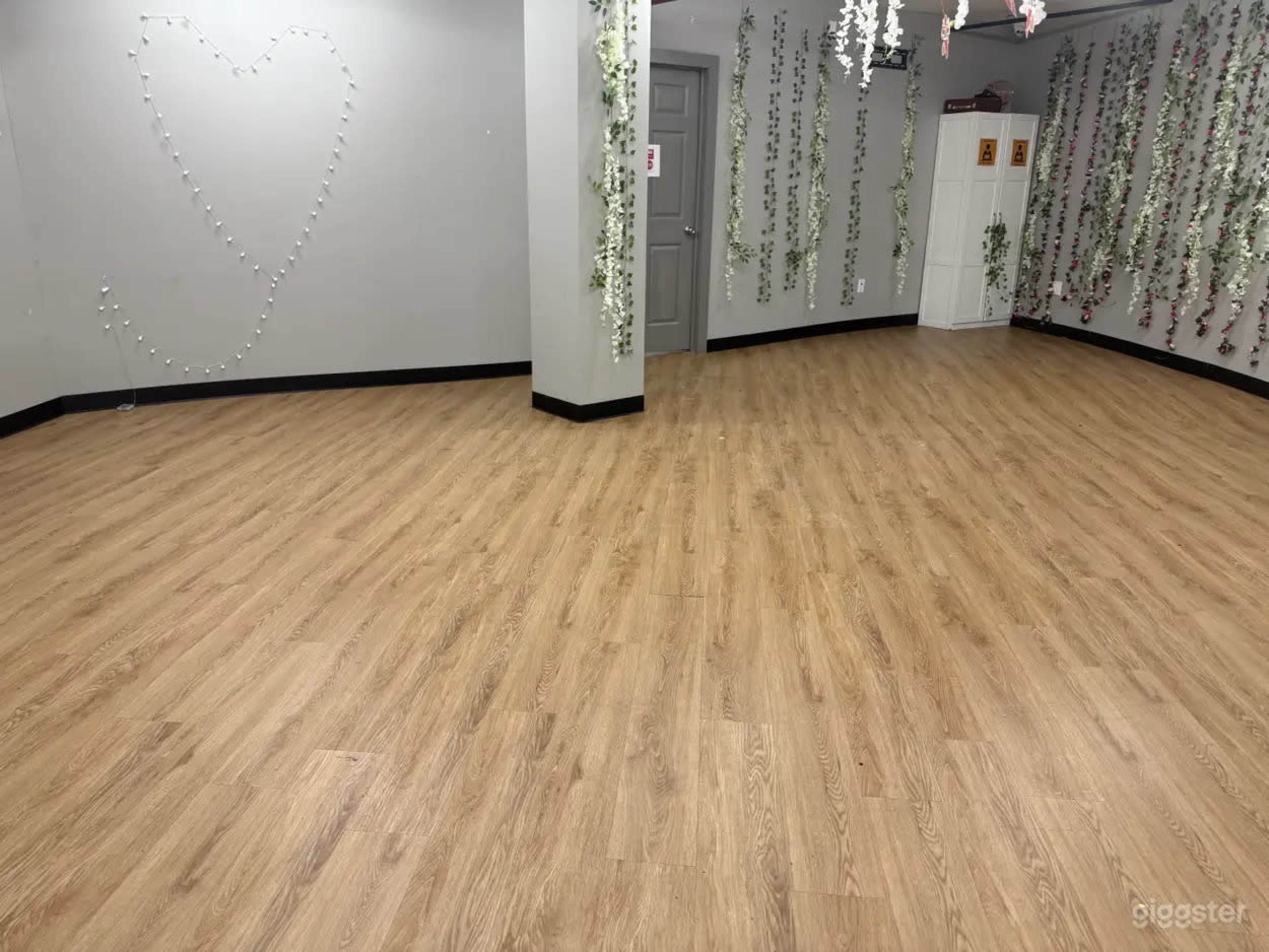 The image shows a spacious room with wooden flooring, decorated with hanging greenery and a heart shape formed by string lights on the wall.