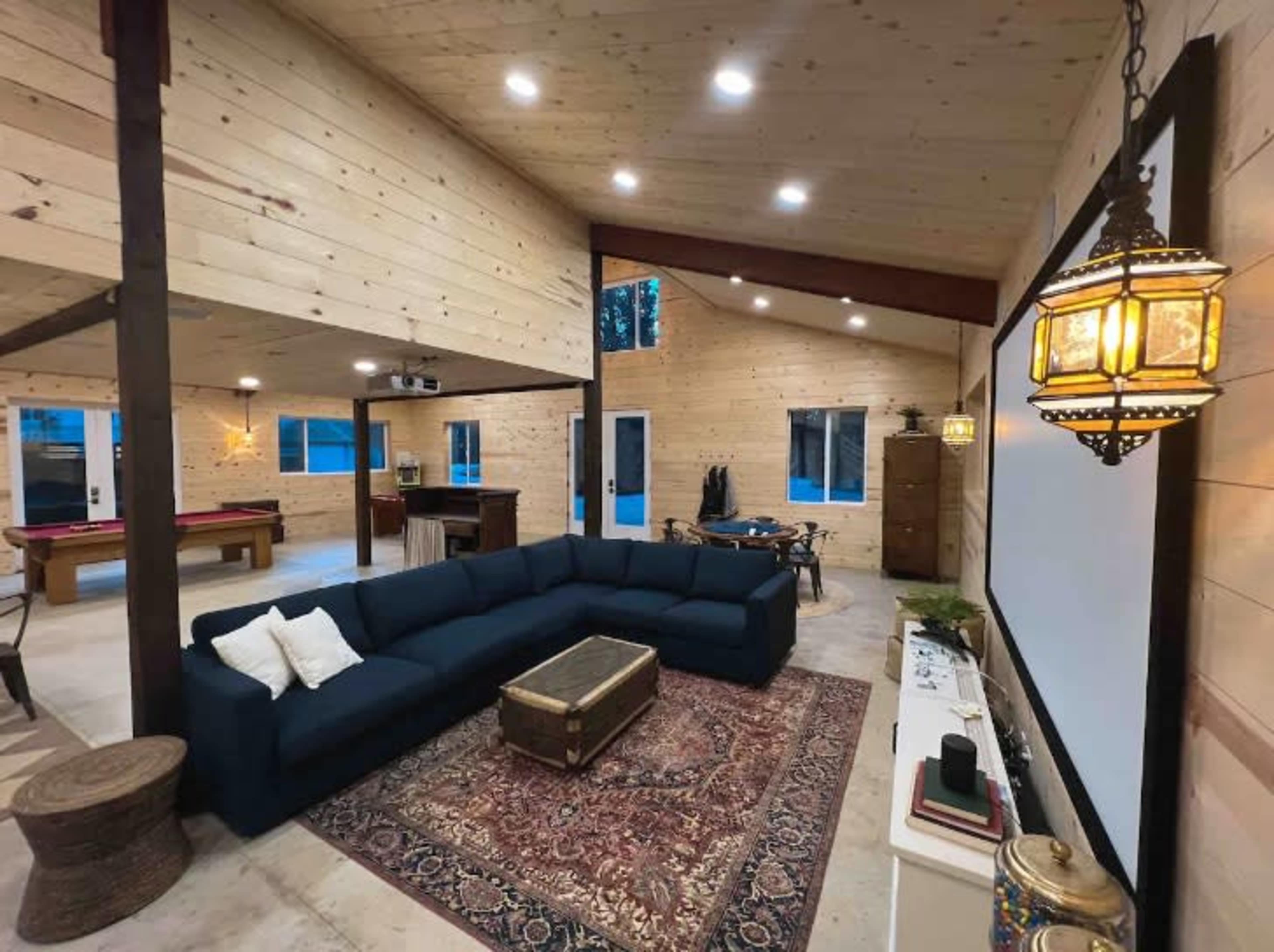 The image shows a spacious interior of a modern wooden-paneled living area with a large sectional sofa, a rug, and a projection screen.