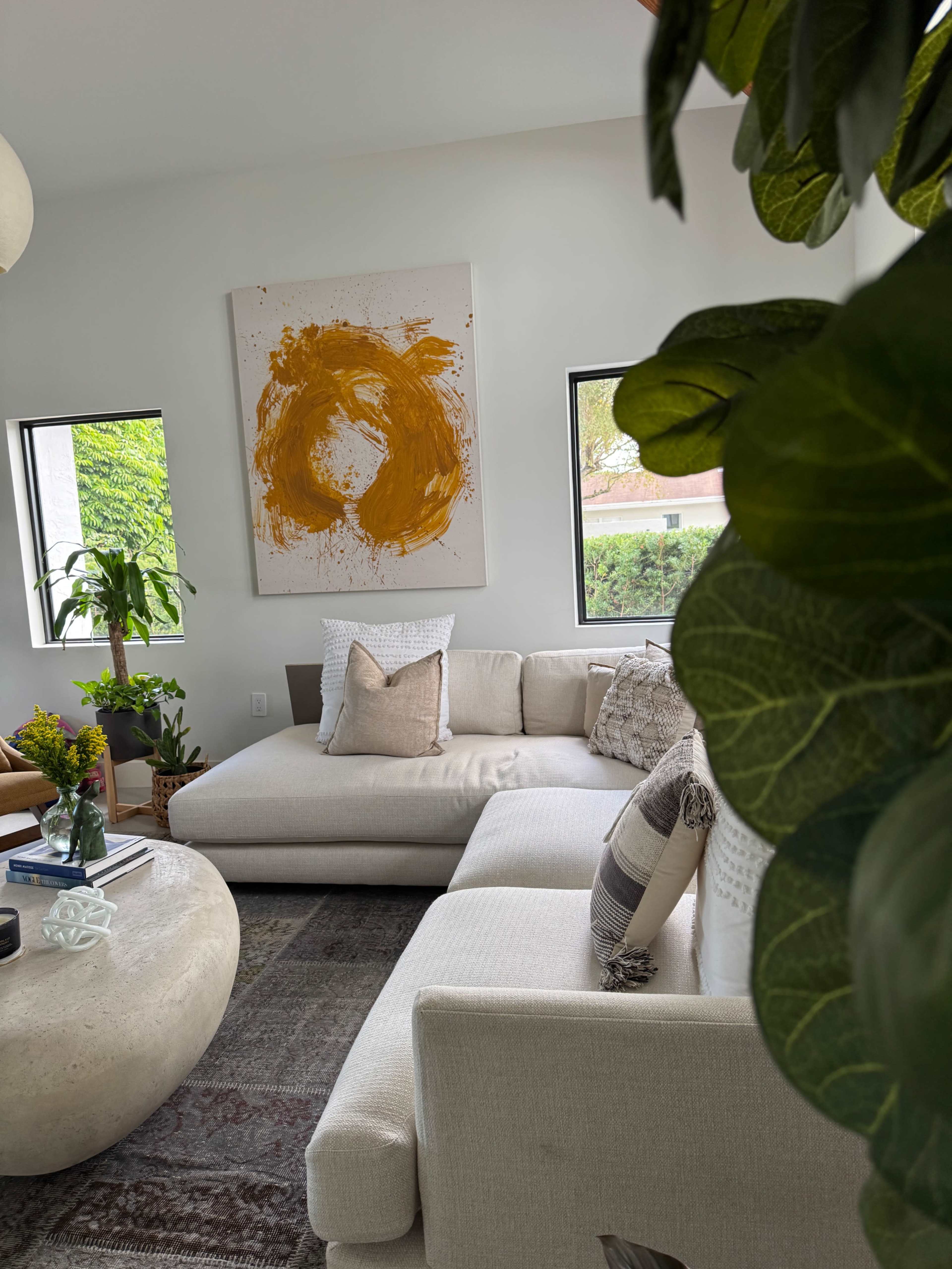 The image shows a cozy living room featuring a large couch, a round coffee table, and a piece of abstract artwork on the wall, surrounded by plants.