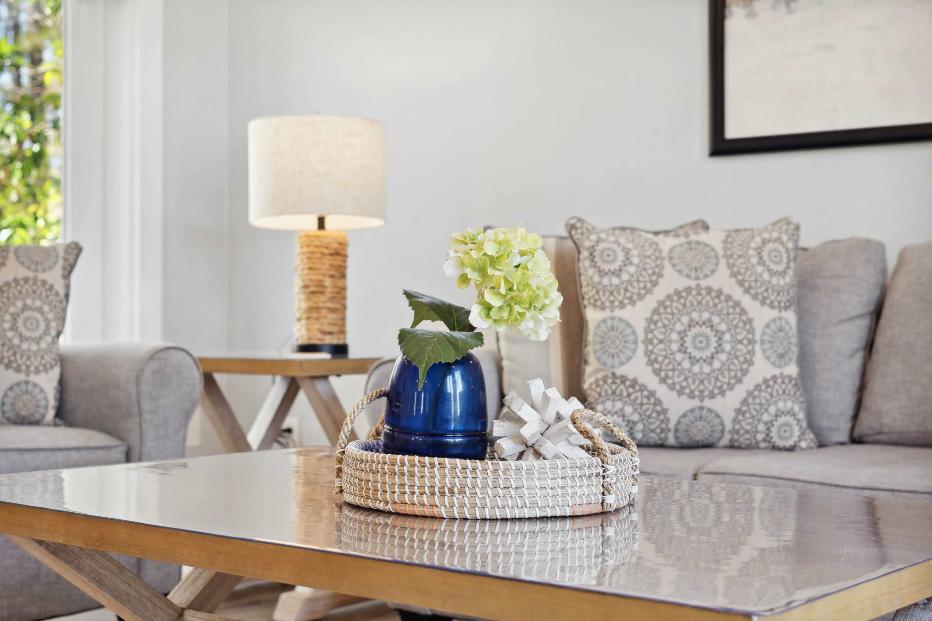A living room features a coffee table with a woven tray holding a blue vase and a white flower, surrounded by gray sofas and a lamp on a side table.