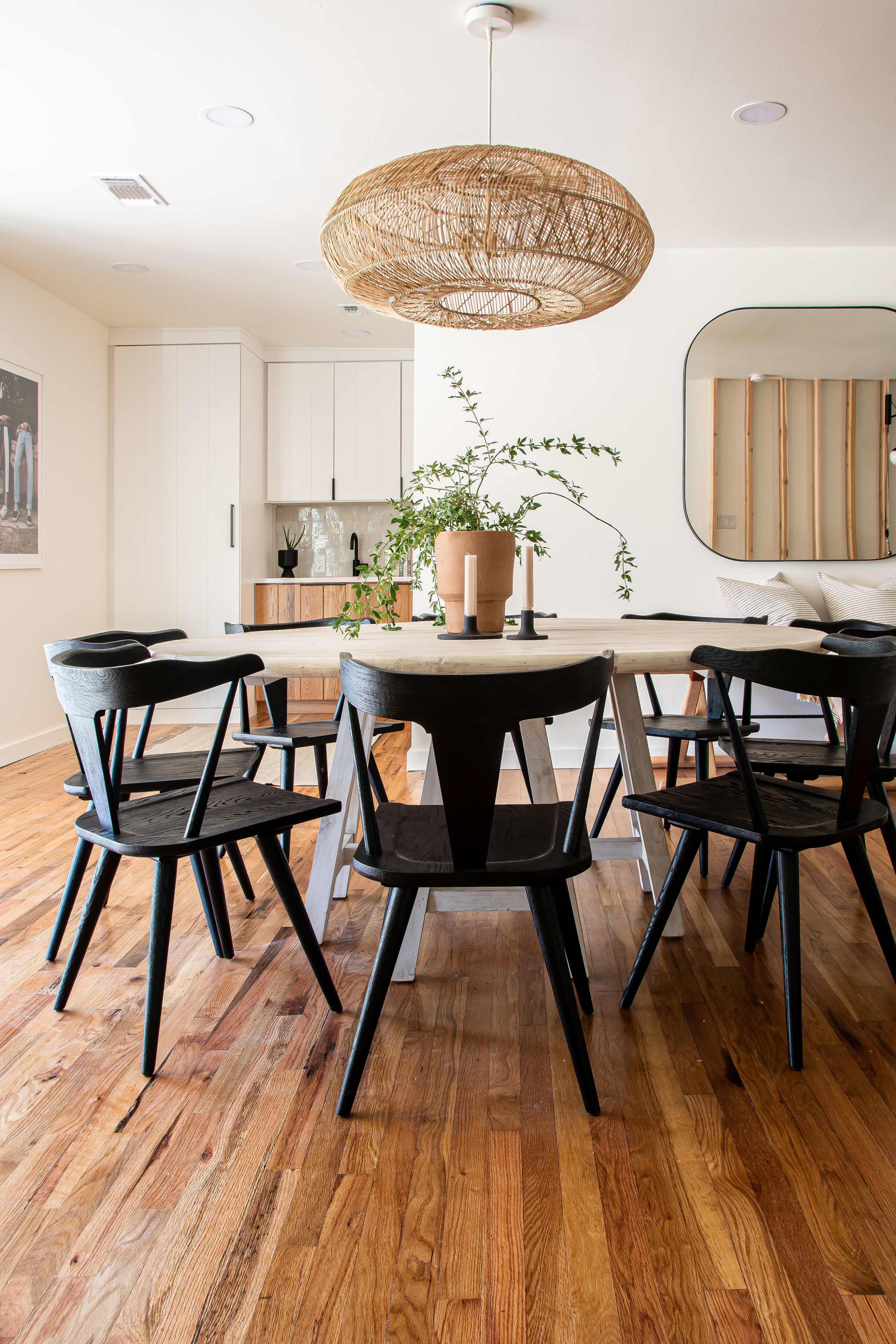 A modern dining room features a round table surrounded by black chairs, with a woven pendant light overhead and a plant in a terracotta pot as a centerpiece.
