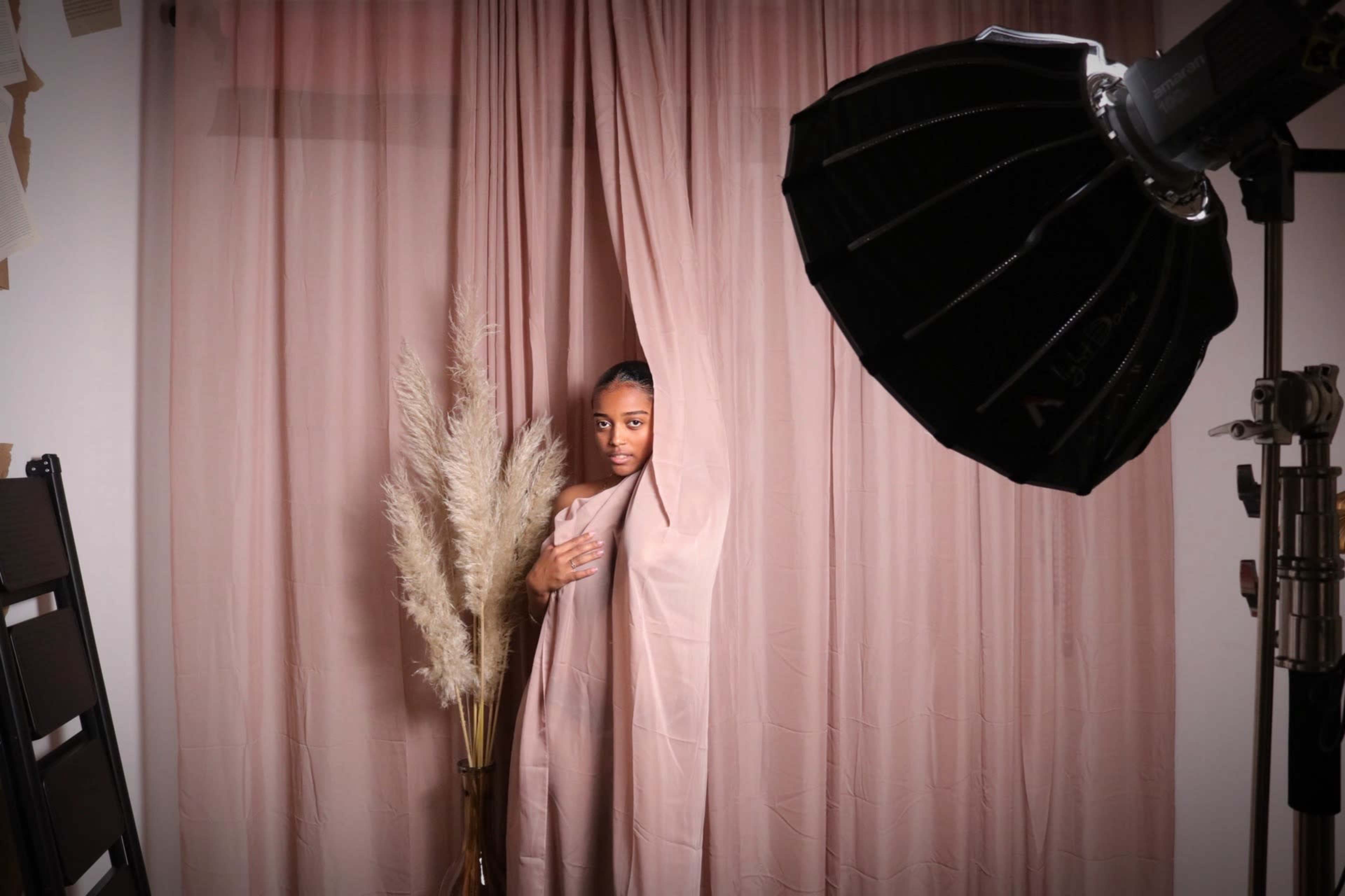 A person stands behind a flowing fabric in front of a soft pink backdrop, with a large photography light source and decorative pampas grass nearby.
